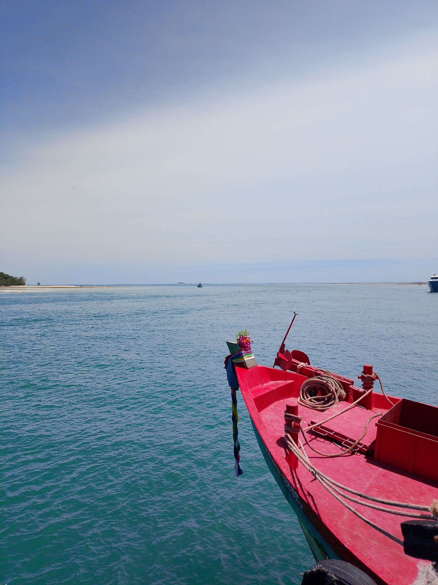 The image captures a bright, sunny day at sea near Koh Phangan, Thailand. In the foreground, the vibrant red bow of a boat is prominently featured, equipped with coiled ropes and a colorful decorative garland tied to its front. The boat is anchored in calm, turquoise water. In the mid-ground, a thin strip of sandy beach stretches across the horizon, with a few scattered trees on the left and a small structure that could be a pier or a lookout point. Further in the distance, another boat is visible on the right, and the sea extends to meet a hazy sky with patches of white clouds. The overall scene suggests a serene coastal environment, likely a harbor or a popular beach area within Koh Phangan.