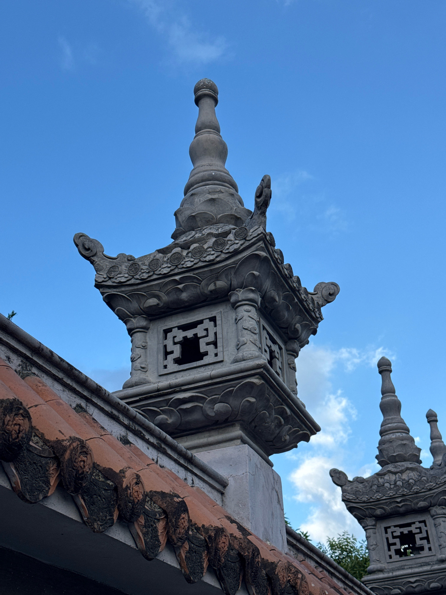 This is a close-up, eye-level, exterior shot of ornate stone pagoda-like structures against a bright blue sky with scattered white clouds. The structures are gray and appear to be made of carved stone with intricate detailing. The foreground is dominated by the top of a red-tiled roof with dark, weathered edges.
In the center of the frame, a detailed stone pagoda spire rises, featuring multiple tiers with decorative carvings and a finial at the very top. To the right, another similar pagoda spire is visible, partially obscured by the first. The architectural style suggests a temple or a historical monument. The environment is clear and bright, with hints of green foliage visible in the background to the right.
The specific location of this image is at the Po Nagar Cham Towers in Nha Trang, Vietnam. These towers are an ancient complex of temples dedicated to the goddess Yan Po Nagar, a significant historical and cultural site in the region.