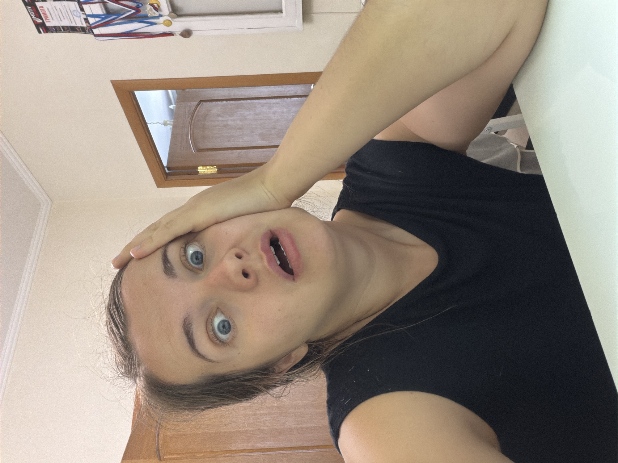 A young woman, appearing to be in her late teens or early twenties, looks directly at the camera with a wide-eyed and open-mouthed expression of surprise or shock. Her light brown hair is pulled back, with some strands framing her face. She wears a dark (likely black) t-shirt, and her right hand is placed on her forehead, enhancing her surprised demeanor.
The scene is indoors, likely a room in a home or apartment in Sochi, Russia. In the background, to the upper left, a white shelf holds several awards, including certificates with red, white, and blue ribbons, and what appear to be gold medals. The Cyrillic word "ГРАМОТА" (GRAMOTA), meaning "Certificate" or "Diploma," is clearly visible on one of the documents. To the right, a wooden door with a light-colored frame and a brass hinge is partially open, revealing a glimpse of another space beyond. A bright, light-colored surface, possibly a table or desk, is visible in the foreground to the right. The lighting is bright and even, consistent with daytime indoor conditions. The overall impression is a candid moment capturing a genuine or playfully exaggerated reaction.