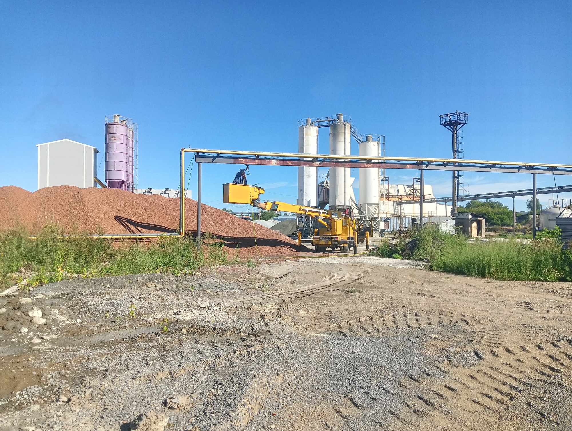 This is an outdoor, wide-angle, eye-level, daylight photograph of an industrial site. The foreground is a rough, gravelly dirt road with tire tracks and some patches of dry soil. In the middle ground, a large pile of reddish-brown gravel is visible to the left. A yellow aerial lift with a man in a blue jacket and black pants in the basket is positioned over a yellow horizontal pipe supported by a metal frame. Further back, several tall, cylindrical industrial silos, some white and some purple, are arranged horizontally across the frame. To the far right, there is a tall, narrow metal tower with a platform at the top. The background is a clear, bright blue sky. The scene appears to be located in a manufacturing or processing facility. There are no discernable landmarks that definitively place this image in Kemerovo, Russia, however, the presence of industrial structures and open land are consistent with industrial zones in such locations.