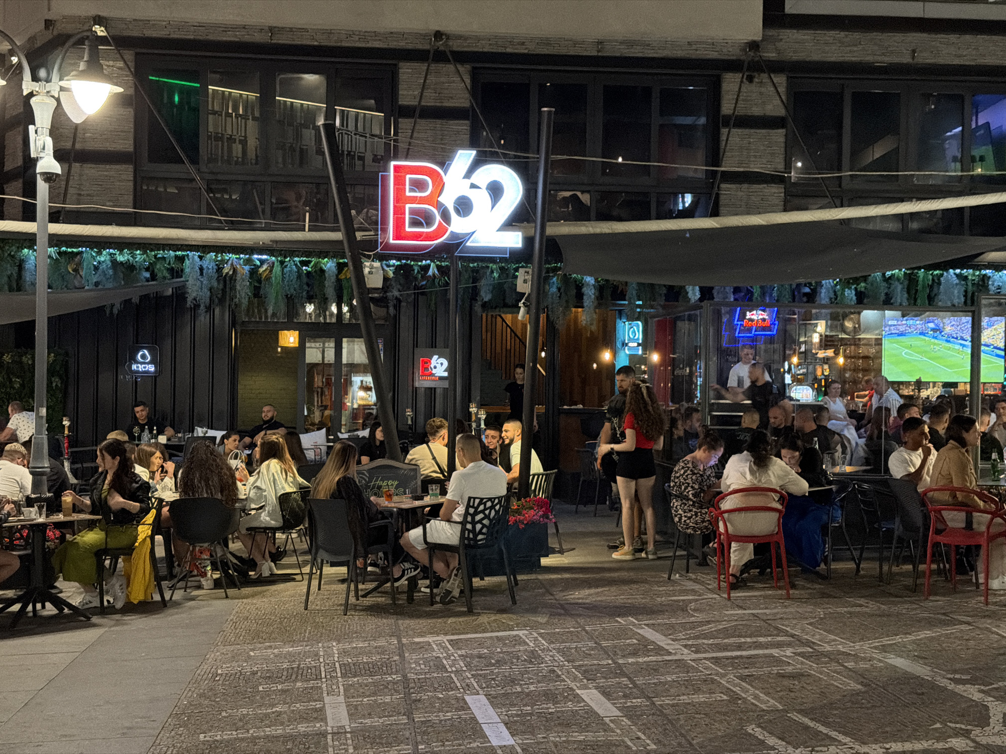 This media file captures an evening scene at an outdoor seating area of a establishment labeled "B62 Lifestyle" in Burgas, Bulgaria. The time of day is clearly night, illuminated by artificial lights from the building's signage, street lamps, and interior lighting.
The primary subjects are groups of people seated at various tables, engaged in conversations, eating, and drinking. The atmosphere appears relaxed and social. A prominent sign with "B62" in red and white neon letters hangs above the entrance, indicating the establishment's name. The building features large windows, some showing interior lighting and patrons. Greenery, possibly artificial hanging plants, adorns the facade above the outdoor seating area.
Visible text includes the "B62 Lifestyle" signage, as well as a smaller "B62" logo on a menu or sign near the entrance. In the background, a large screen displays a soccer match. The ground is paved with patterned tiles. The weather appears to be clear, and the presence of people dining outdoors suggests mild temperatures. The overall impression is a lively evening at a popular spot.