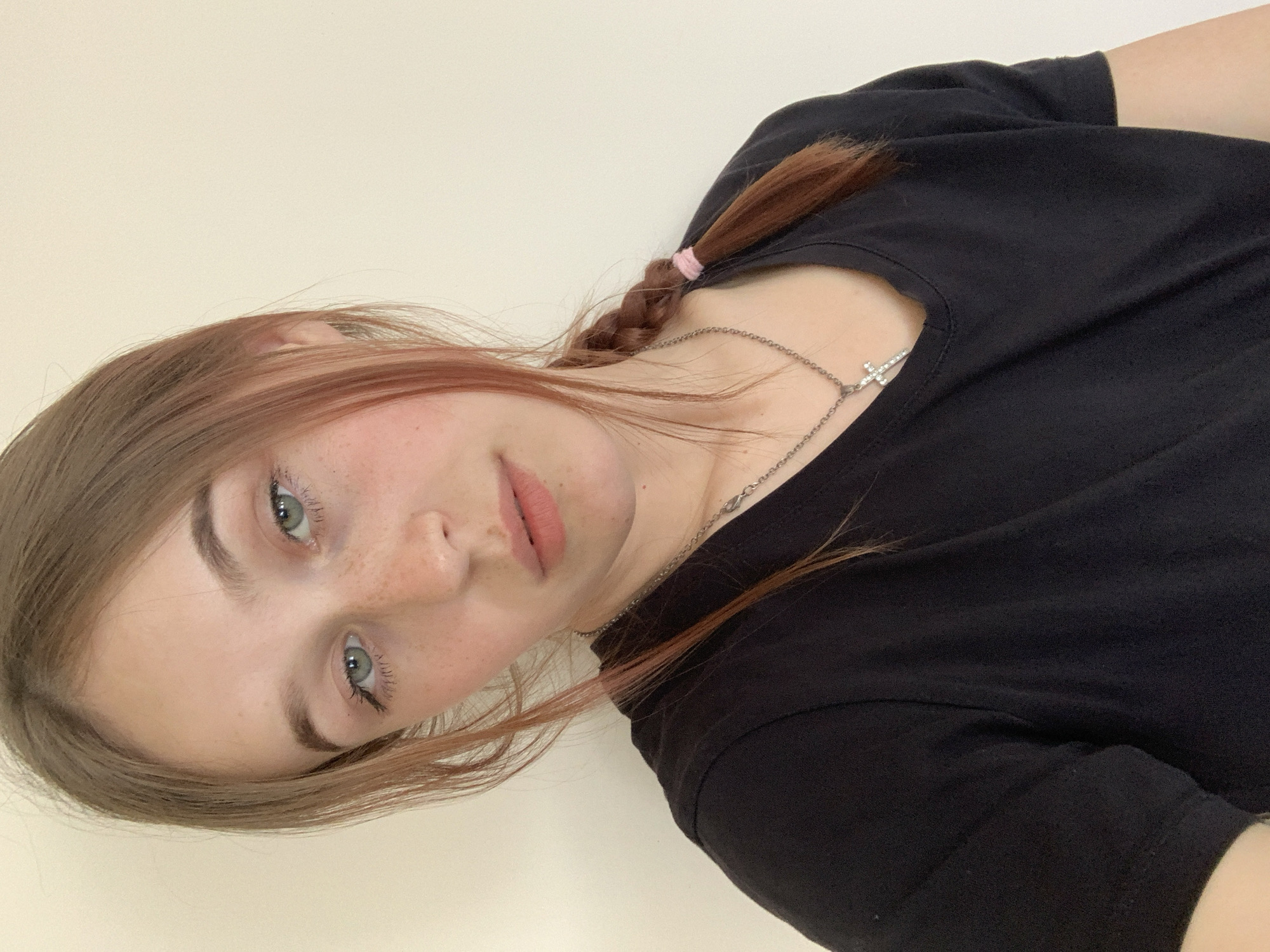 A young woman with light brown hair and freckles poses for a selfie against a light beige wall. She is wearing a black t-shirt and a silver necklace with a cross pendant. Part of her hair is braided and secured with a pink hair tie. The location is unknown and unrelated to Фоки, Russia.