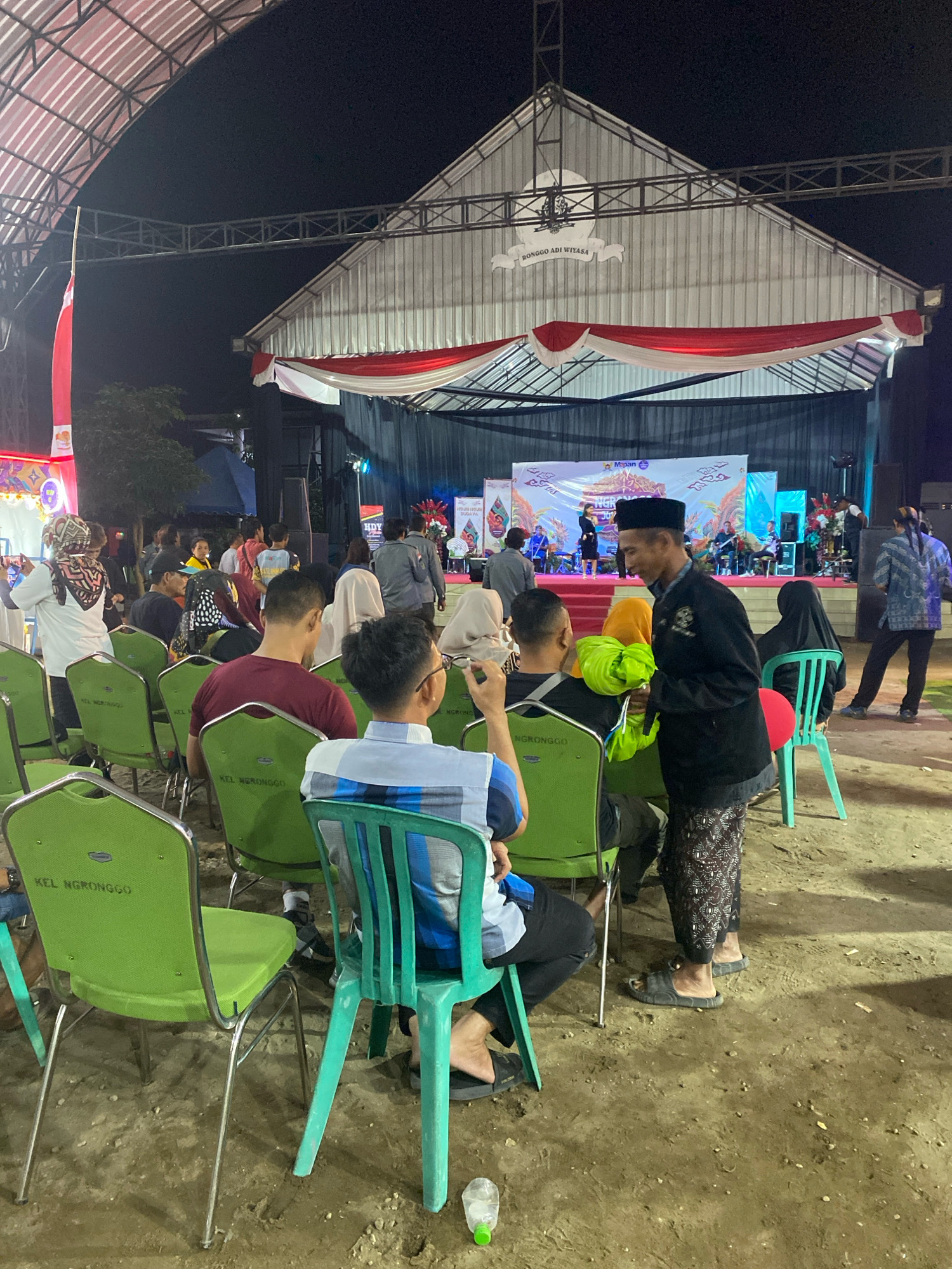 This image captures an outdoor evening event likely taking place in the city of Kediri, Indonesia, possibly at a community gathering or cultural performance. In the foreground, several people are seated on green plastic chairs arranged in rows. The chairs have metal frames and backs with the text "KEL NGRONGGO" printed on them. The ground is dirt and appears to be uneven.
In the mid-ground, more people are visible, some standing and others seated. A man in a black cap and black attire, possibly a uniform or traditional clothing, is interacting with someone, holding a bright green object. Further back, a stage is set up with a backdrop featuring graphics and text. Performers are visible on the stage, with musical instruments and lighting equipment present. A large banner is hung above the stage with Indonesian text.
The background shows a structure with a pitched roof, decorated with red and white draped fabric, suggesting a festive or official occasion. The lighting is artificial, typical for evening events. The overall scene depicts a community gathering in an open-air venue, possibly in a sub-district or village area of Kediri, given the "KEL NGRONGGO" text on the chairs, which likely refers to a local administrative unit.