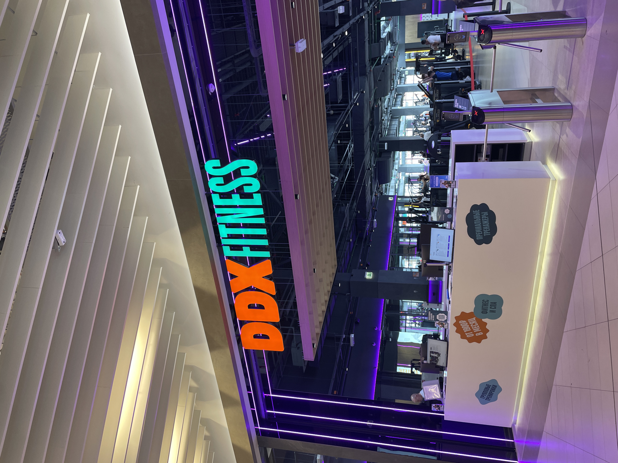 The image depicts the interior of the DDX Fitness gym in Kazan, Russia. A brightly lit, orange and green DDX Fitness sign is visible. The gym has a high ceiling with exposed ductwork and structural elements. Rows of exercise equipment are seen, with people using the machines. A white reception desk with blue and orange signage is present, and an employee is visible behind it. There are turnstiles for entry. The lighting is a mix of white and purple neon.