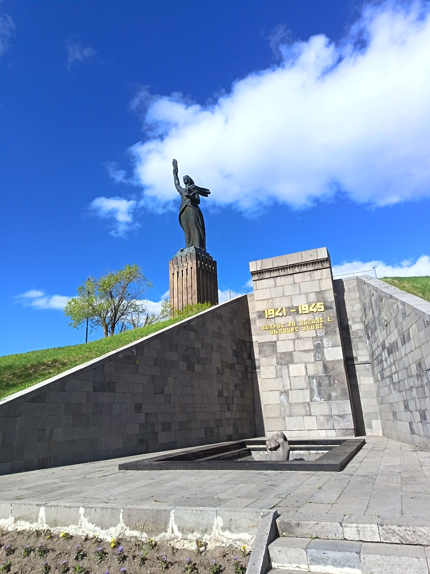 The image shows the Mother Armenia statue and memorial complex in Yerevan, Armenia, under a blue sky with scattered white clouds. The statue, located atop a tall pedestal, depicts a woman holding a sword above her head, symbolizing victory. To the right of the statue and slightly in front is a monument with the dates "1941-1945" inscribed in gold lettering, likely commemorating World War II. Beneath the dates, there is additional text in a non-English script.
In front of the monument, there is a stone structure, possibly a fountain or a memorial, with a hand emerging from the center. The entire complex is constructed with gray stone blocks, giving it a somber and monumental appearance. The area surrounding the complex features greenery and a few small flowers at the base of the structure. The image is taken from a low angle, emphasizing the height and grandeur of the statue and memorial.