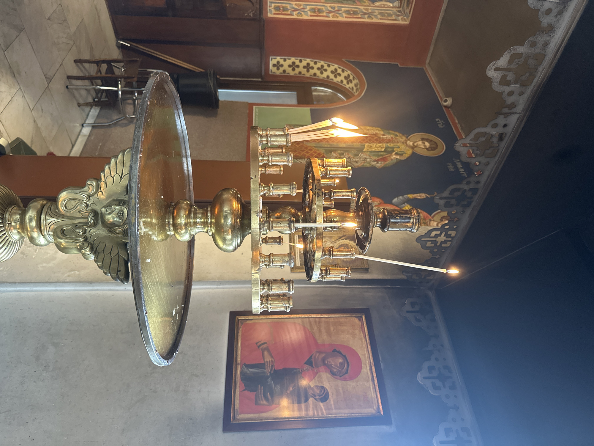 The image captures the interior of an Eastern Orthodox church or monastery, likely in Sofia, Bulgaria, characterized by rich religious iconography and warm lighting.
In the foreground stands a magnificent, multi-tiered brass candelabrum, a votive candle stand, adorned with intricate carvings, including an angelic or eagle-like figure at its base. Numerous thin white candles are placed in its holders, several of which are actively burning, casting bright, warm flames and contributing to the sacred atmosphere.
The background reveals ornate frescoes on the upper walls and ceiling. One prominent fresco depicts a haloed figure, identifiable as "Saint John" ("О АГІОС ІΩАННН" in Greek script) based on the visible text above and below the halo. This figure is robed in green and gold, rendered in the traditional Byzantine style. Decorative patterns, featuring cross-like motifs and geometric designs, frame the frescoes.
On a lower wall, a framed icon is displayed, depicting a Madonna and Child-like figure. The Madonna is portrayed with a red head covering and robe, holding a child figure. The artwork appears to be in a traditional Eastern Orthodox style.
The floor on the left side of the image is tiled. In the deeper background, there are stacked wooden chairs and other dark furniture, suggesting areas for seating or utility within the religious space. The overall lighting is dim, with the candelabrum's flames providing significant illumination, creating a reverent and contemplative mood. No people are visible, emphasizing the quiet sanctity of the space.