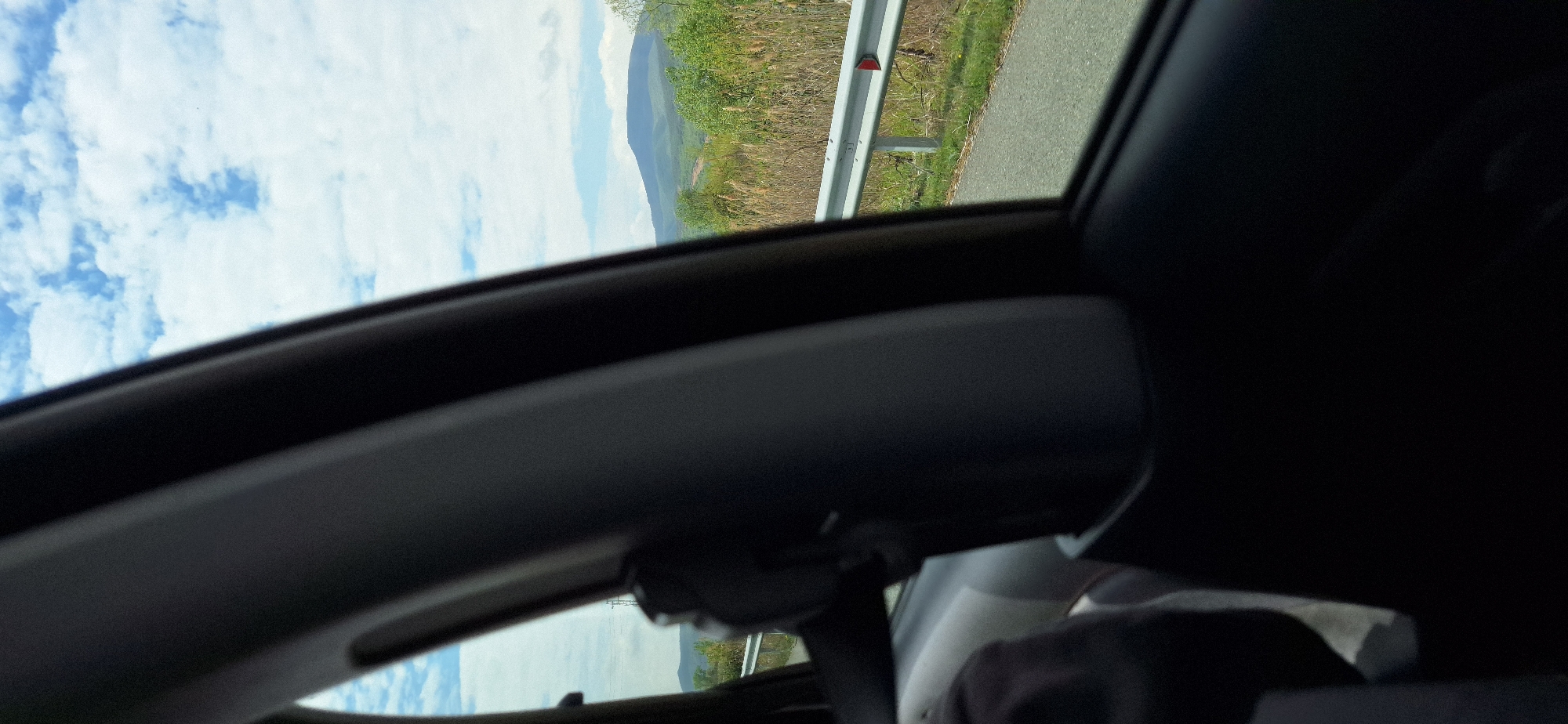 The image shows a view from inside a car, possibly a sunroof perspective. Outside the car, there is a roadside with a guardrail, vegetation, and a distant mountain. The sky is visible with clouds. There is no indication of being in the city of Valkovo, Bulgaria. There are no visible people.