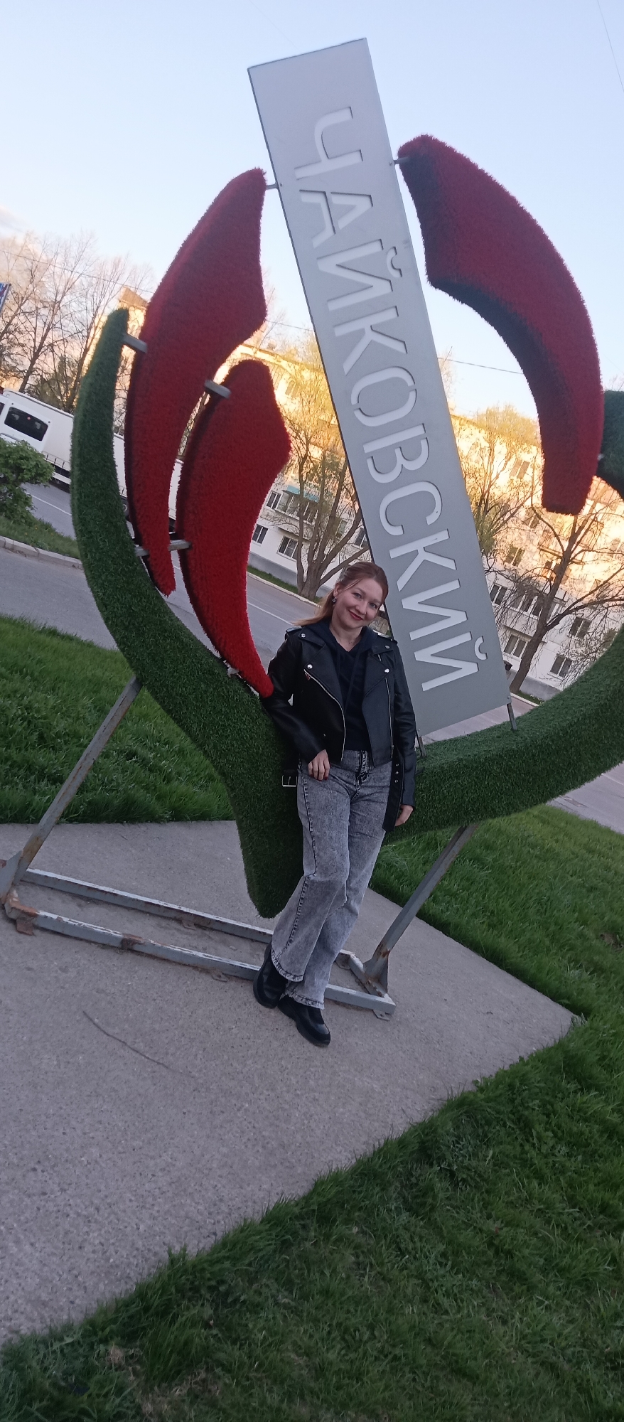 A full shot shows a woman posing in front of the "Чайковский" (Tchaikovsky) sign in the city of Tchaikovsky. The sign is made of green and red topiary in a stylized tulip shape, with a gray metal sign displaying the city's name in Cyrillic. The woman is wearing a black leather jacket, black shirt, acid wash jeans, and black boots. She is leaning against the bottom of the flower structure. Behind her is a street with a bus and a building with visible windows. The surrounding area is green grass and a concrete sidewalk. The lighting suggests it is either early morning or late afternoon.