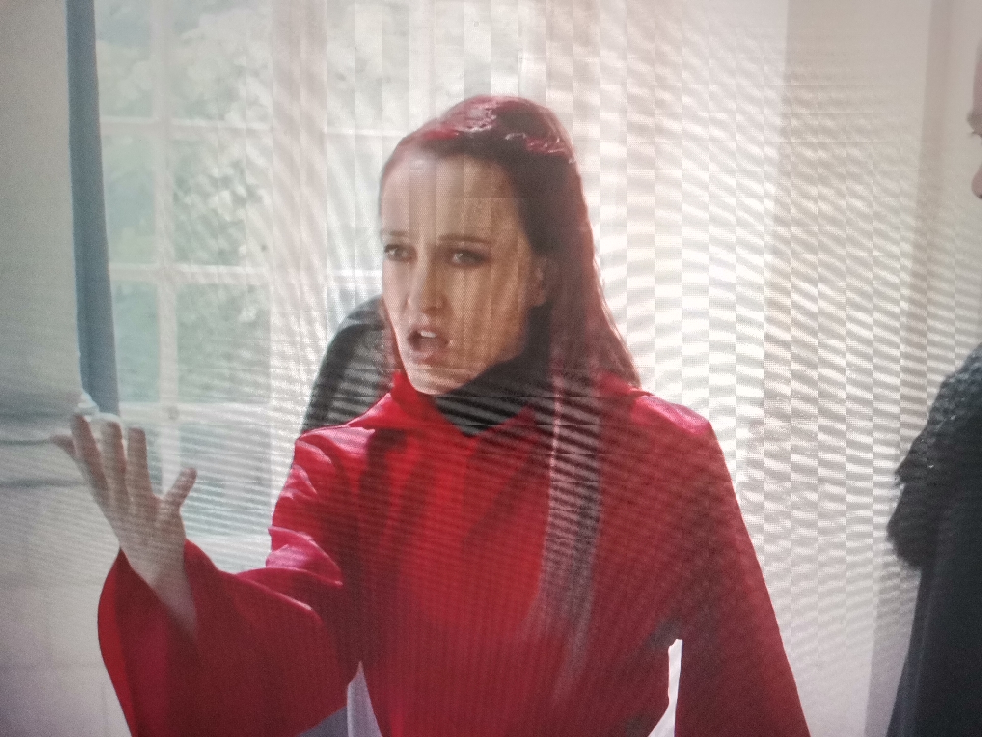 A woman with long, red hair pulled back, wearing a red robe over a black turtleneck, gestures with her left hand while speaking. Her mouth is open, and she appears animated. She stands indoors, near a window with large panes showing greenery outside. Another person in dark clothing is partially visible to her right. The setting appears to be a building with architectural details suggesting it could be a historical site, possibly located in the city of Belaazyorsk, Belarus. The lighting is bright, coming from the window and possibly overhead.