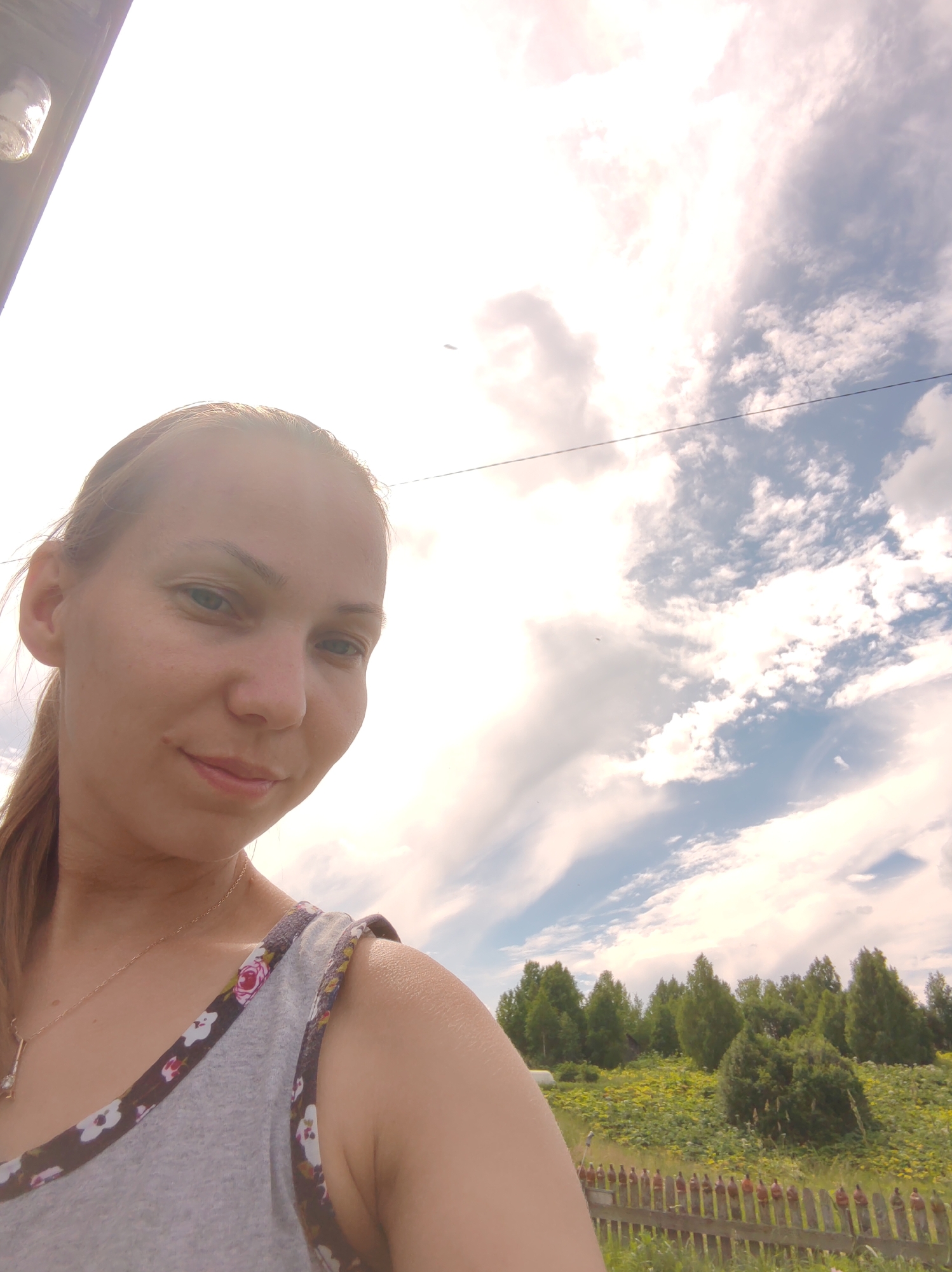 The image is a selfie taken by a woman outdoors in a rural setting, likely on a sunny day with a bright sky and scattered clouds. The woman, who appears to be of East Asian descent, is in the foreground, her face and upper body visible. She has light brown hair pulled back and is wearing a gray tank top with a floral trim. She is looking towards the camera with a slight smile. Behind her, a landscape is visible with green trees and foliage, a wooden fence, and a bright, overcast sky. The location is specified as Chukhlem, Russia. The overall scene suggests a casual outdoor moment.