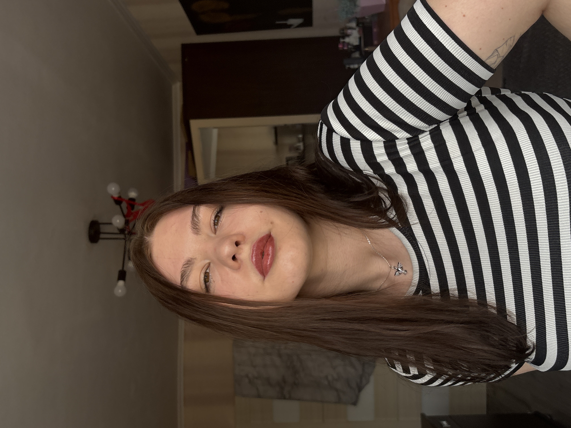 A woman with long brown hair is facing the camera. She wears a black and white striped top and a silver necklace with a pendant. The background shows a room interior with a lamp and a door. The location is the city of Perm, Russia.