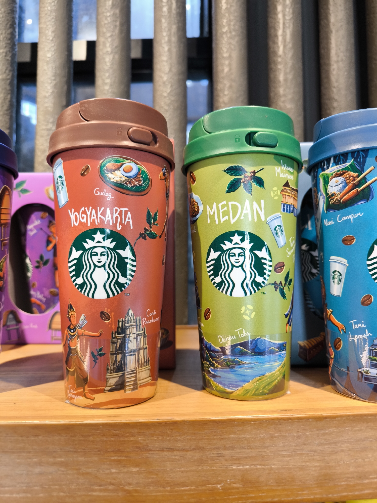 The image shows a close-up of three Starbucks tumblers, each depicting scenes and named after different Indonesian cities: Yogyakarta, Medan, and an unreadable city. The Yogyakarta tumbler is orange and shows images of Gudeg, a traditional dish, and Candi Prambanan, a temple. The Medan tumbler is green and displays Istana Maimoon, a palace, and Danau Toba, a lake. The tumblers sit on a wooden shelf in front of a background of grey vertical bars. The location is not explicitly stated as Kuta, Indonesia, but the cities depicted are located in Indonesia.