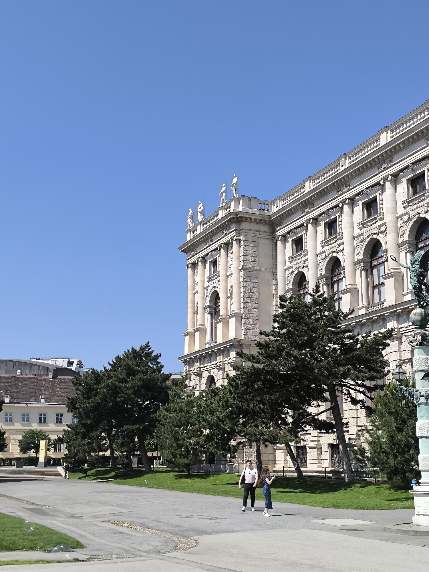 The image shows a daytime scene at the Museumsplatz in Vienna, Austria. The Kunsthistorisches Museum (Museum of Art History) occupies a large portion of the frame, displaying its ornate facade with sculptures along the roofline and detailed architectural elements.
Two individuals, a man and a woman, are walking on a paved area in front of the museum. The man is wearing dark trousers, a light-colored shirt, and sunglasses. The woman is wearing a blue patterned dress.
Trees are positioned between the paved area and the museum, partially obscuring the lower part of the building. A grassy area is visible behind the trees.
The sky is clear and blue. In the distance, another building is visible. The foreground includes pavement with some scattered litter. A statue or monument is partially visible on the right side of the frame.