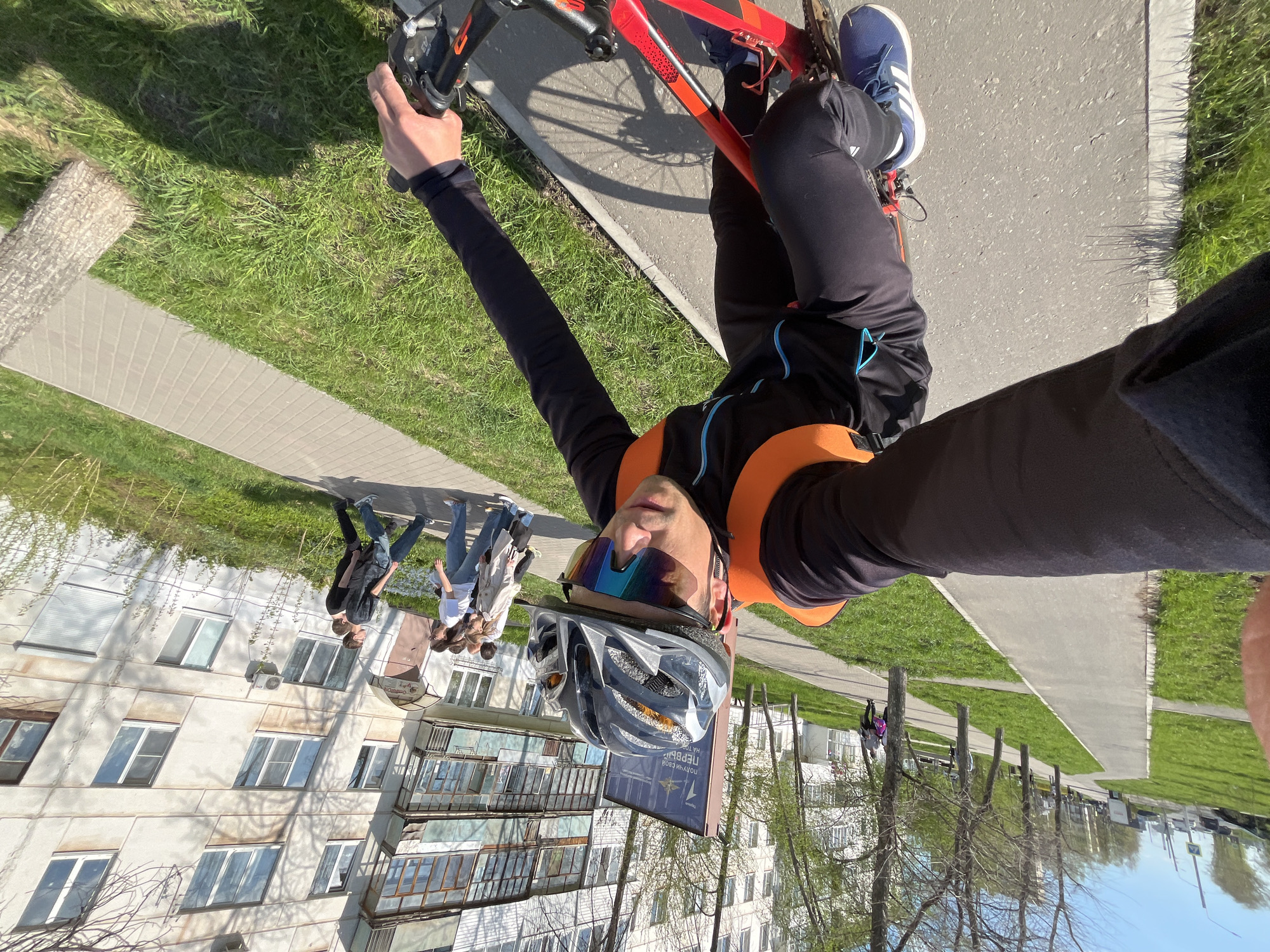 The image shows a man on a bicycle taking a selfie. He is wearing a helmet, sunglasses, and athletic clothing. The bicycle is red and black. The background includes a building with several windows and balconies, a group of people walking on a sidewalk, grass, trees, and a paved path. The building appears to be a multi-story apartment complex with a sign visible. Based on the architecture and visible Cyrillic text, the location is likely Chaikovsky, Russia.
