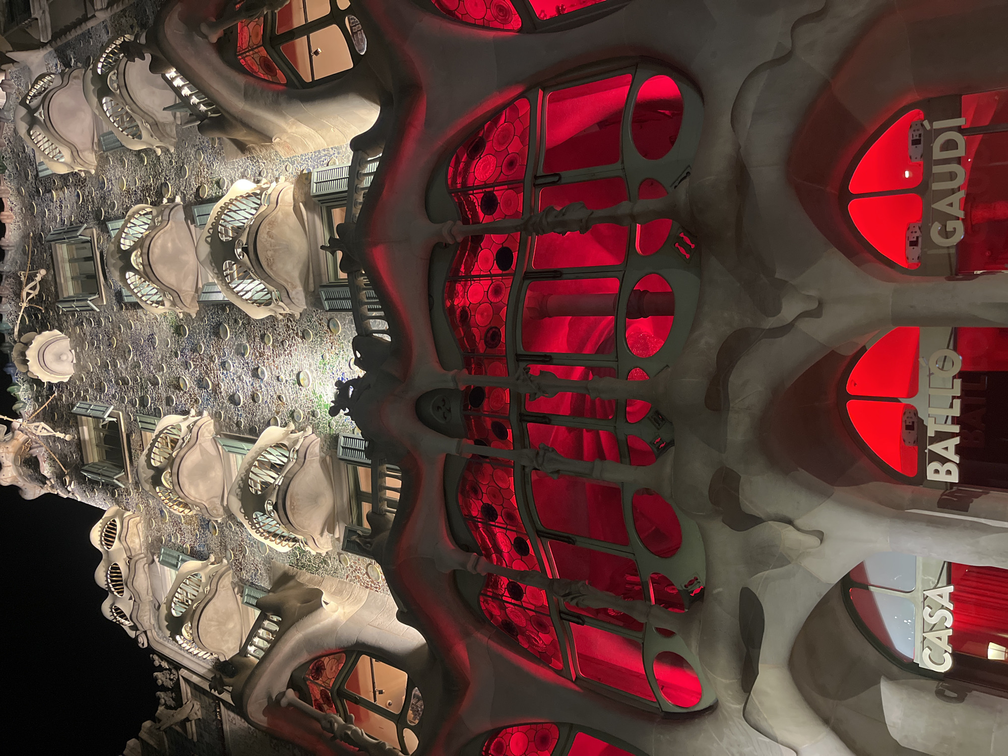 The image captures a nighttime view of Casa Batlló, a modernist building located on Passeig de Gràcia in Barcelona, Spain. The facade is illuminated with red lights, casting a vibrant glow on the organic, bone-like architectural elements. The upper floors feature distinct balconies with skeletal, mask-like shapes and rounded windows, while the lower levels display large windows with red stained glass, creating a dramatic and ethereal effect. The text "CASA", "BATLLÓ", and "GAUDÍ" are visible in illuminated white lettering on the lower facade. The sky above is dark, indicating it is nighttime. The building's unique architectural style, characterized by its undulating forms and mosaic facade, is a prominent feature.