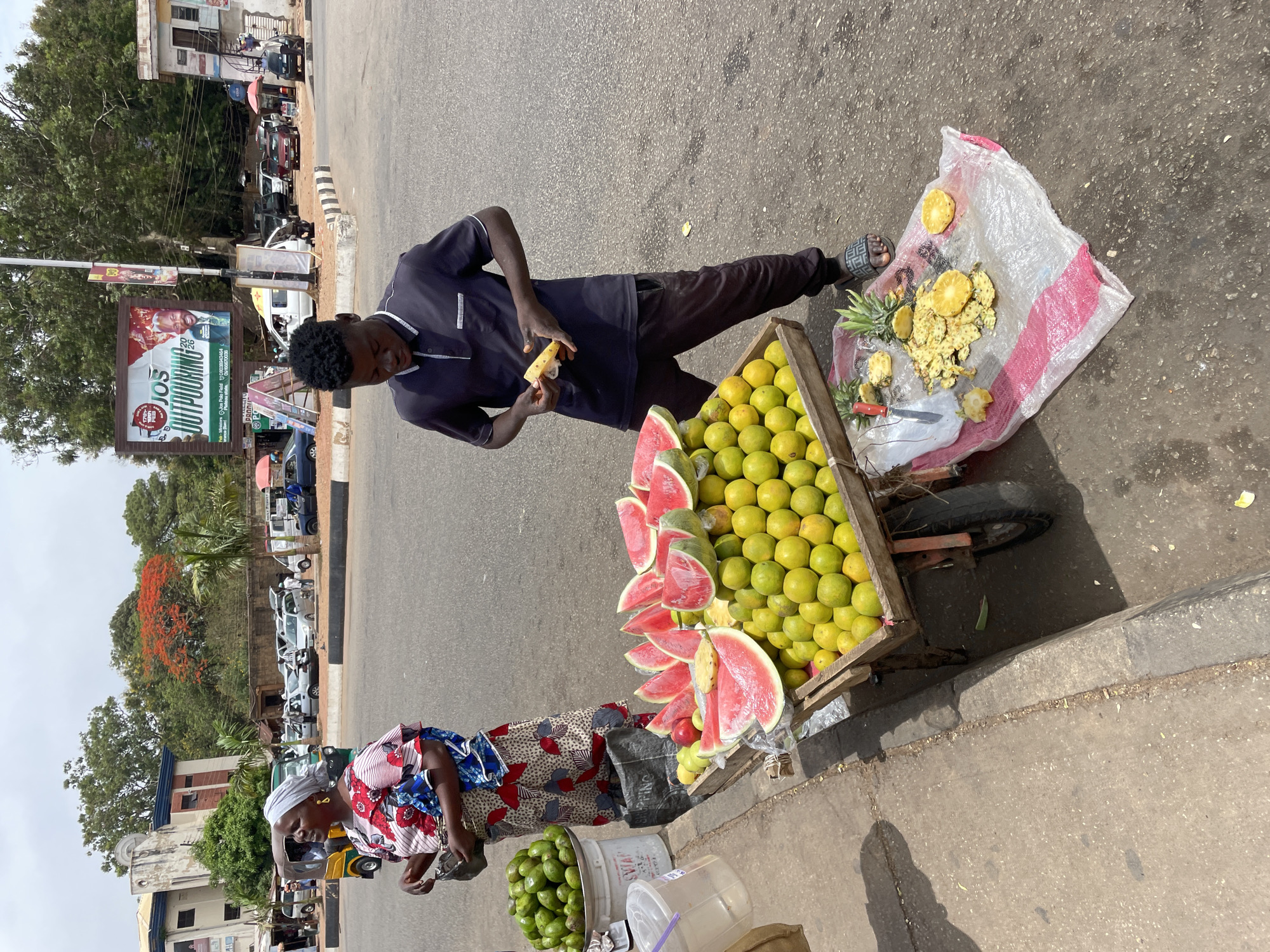 This image captures a vibrant street scene, likely a fruit vendor's stall, in Jos, Nigeria, during daylight hours, indicated by the bright lighting and clear shadows.
In the foreground, a young man, dressed in a dark blue polo shirt and dark trousers, stands behind a wooden fruit cart. He has short, dark curly hair and is holding a piece of fruit, possibly a pineapple slice, appearing to be preparing or eating it. His sandals feature a distinctive pattern.
The fruit cart is laden with an abundance of fresh produce. One section holds numerous bright green-yellow citrus fruits, likely oranges or limes. Adjacent to these are several large, bright red watermelon slices, neatly arranged. On the ground next to the cart, a white plastic bag contains discarded pineapple peels and some prepared pineapple slices, along with a small knife, suggesting on-the-spot fruit preparation.
Standing further back and to the left of the cart is a woman wearing a colorful patterned dress and a light-colored headscarf. She appears to be observing or attending to the stall, with her hands clasped in front of her. Near her, a white bucket is filled with green fruits, possibly mangoes or avocados, and other clear plastic containers are visible on the ground.
The background reveals a bustling street with a paved road, a curb, and buildings lining the street. Various vehicles, including cars and a green tricycle (keke napep), are parked or moving along the road. Overhead, large trees provide shade, and a tree with striking red blossoms adds a splash of color. A prominent billboard in the background clearly displays the text "JOS OUTPOURING" along with contact details and location "Plateau Hotel." Other smaller, less legible advertisements are also visible. The overall scene depicts a typical, busy urban environment centered around a fresh fruit market stall.