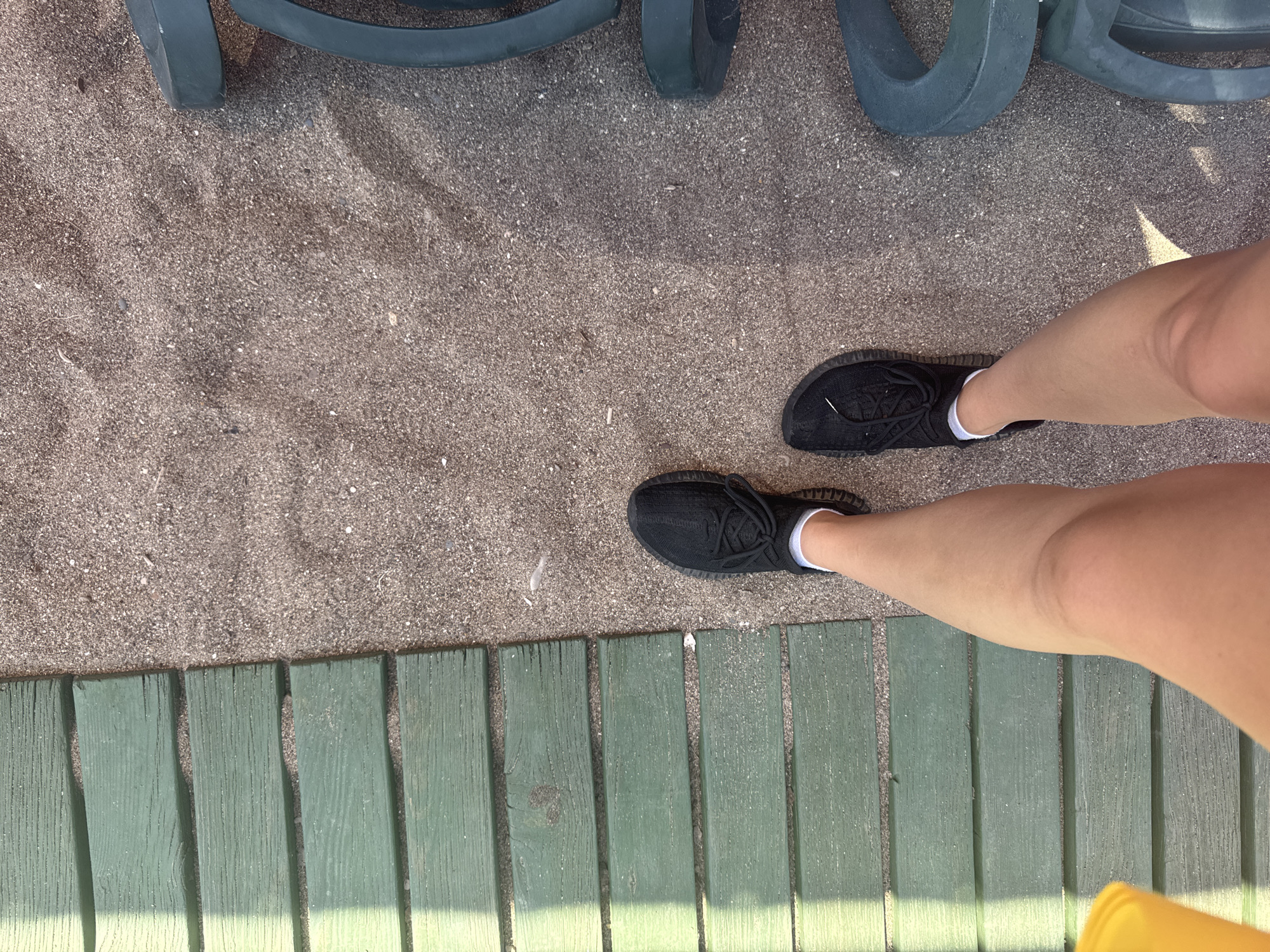 A high-angle, medium shot captures a person's legs standing on a sandy area next to a green wooden walkway. The person is wearing black athletic shoes with white socks. In the background, part of a green plastic lounge chair is visible. The scene is outdoors, likely in a beachside or park setting in Aksu, Turkey. The lighting suggests it is daytime, with shadows cast across the wooden walkway.