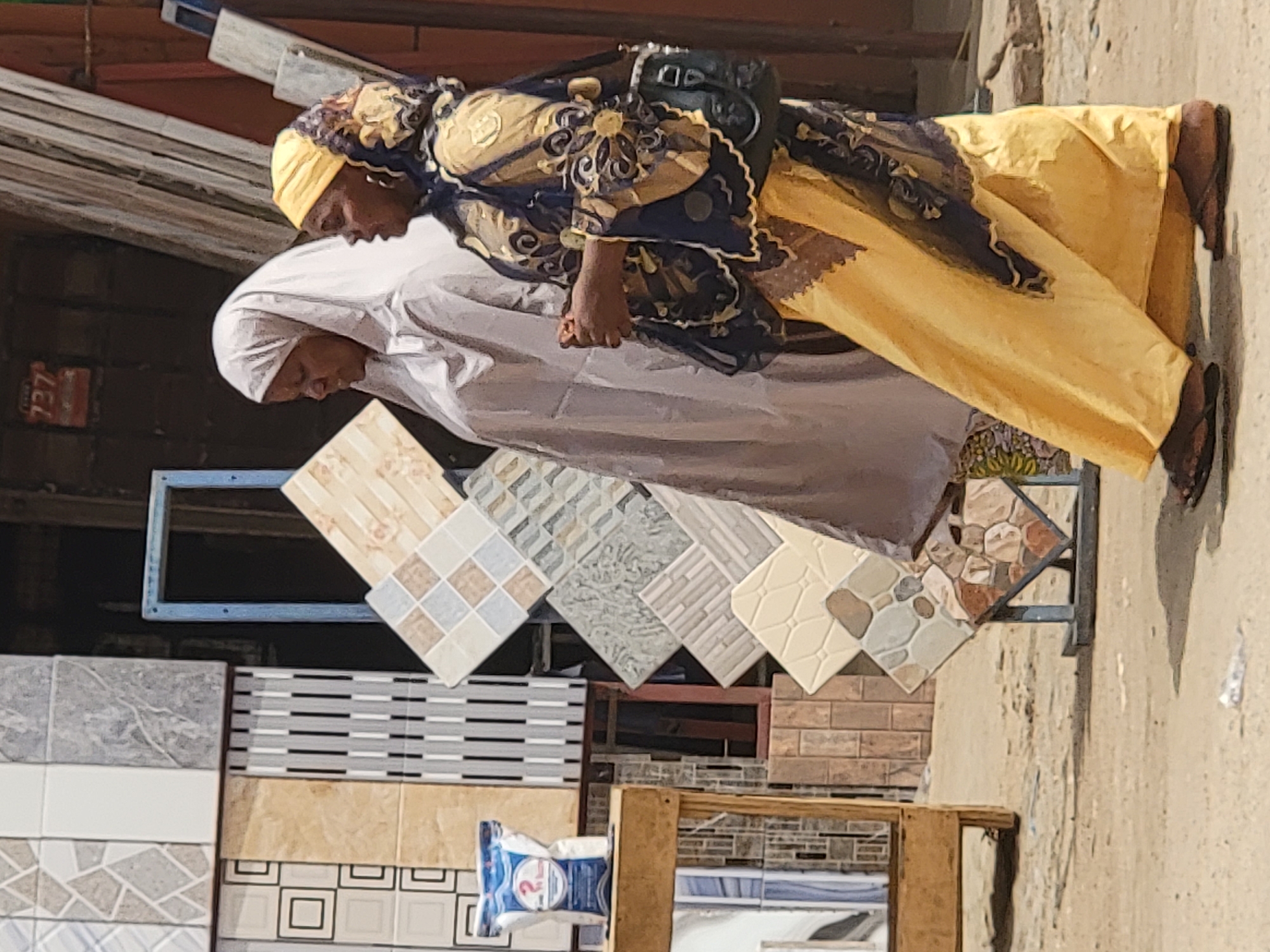 The image, rotated 90 degrees clockwise, depicts a street scene in Maiburiji, Nigeria, featuring two women examining a display of decorative tiles.
The central figures are two women with dark complexions. The woman on the right wears a bright yellow head wrap and an elaborate dark blue and gold patterned top over a flowing yellow dress. A dark handbag hangs from her shoulder, and she wears dark flip-flops. The woman on the left is dressed in a simple white head covering and a loose white garment. Both women are looking downwards, seemingly focused on the tile display in front of them, suggesting they are browsing or selecting items. Their expressions appear calm and concentrated.
The setting is an outdoor shop or market stall showcasing a variety of floor and wall tiles. Numerous square and rectangular tiles are arranged on metal stands and racks, some at an angle. These tiles feature diverse patterns, colors (including beige, white, grey, brown, and black), and textures, some with geometric designs, others mimicking brick or natural stone.
In the background, on the far left, a red rectangular object is visible, bearing the numbers "737". Near the bottom center, a white plastic bag with a blue label clearly shows the number "2". The ground is a light-colored, uneven surface, likely dirt or concrete. The scene is illuminated by bright natural light, indicating it is daytime. The overall impression is that of everyday commerce in a bustling local market environment.