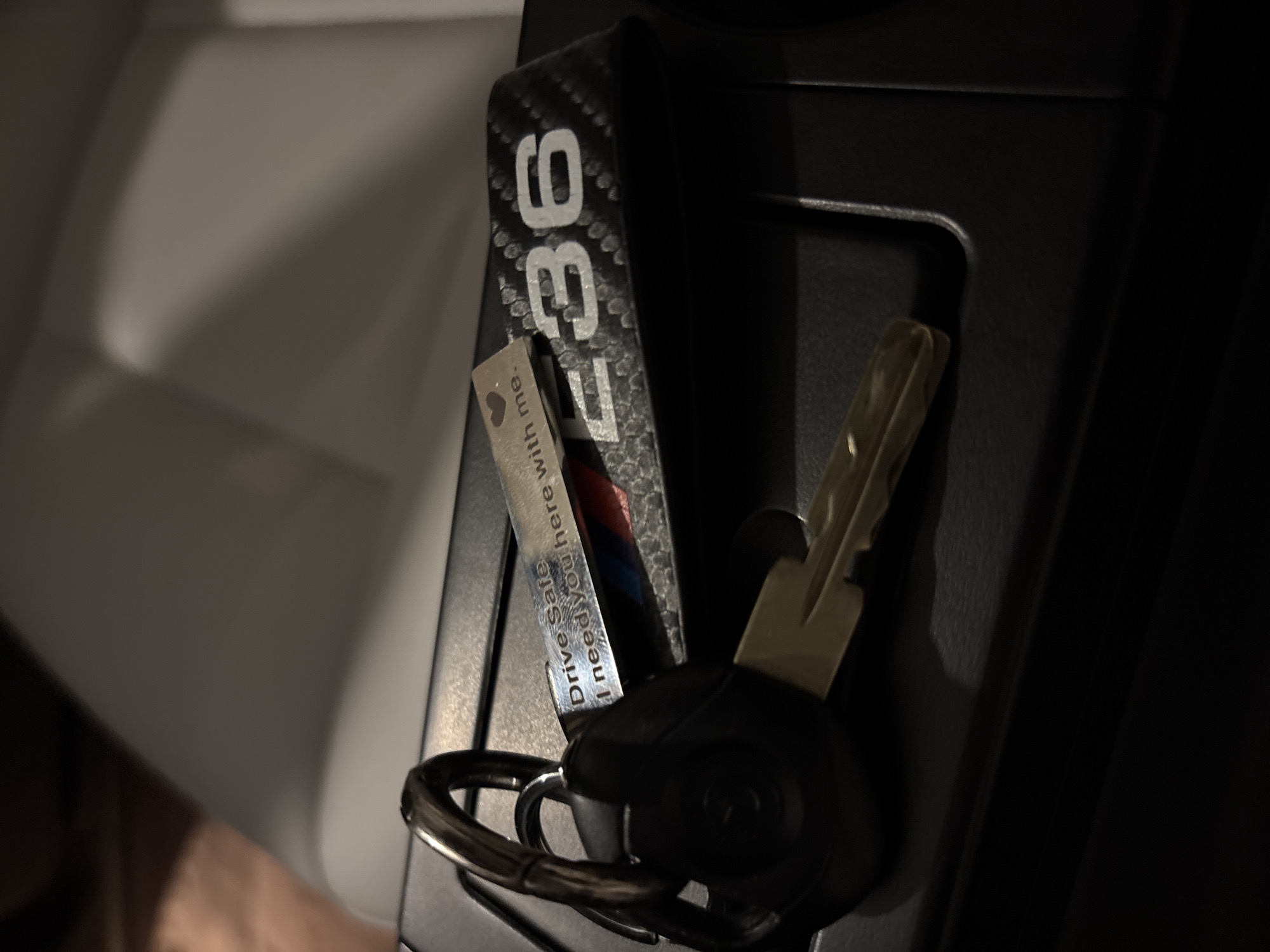 The image shows a set of car keys with a keychain that says "E36". The keychain is attached to a keyring with the car key and a small tag. The tag reads "Drive Safe. I need you here with me." The keys and keychain are resting in the car's center console. The environment is dark, suggesting the interior of a car. The location is Unknown, Unknown. There are no people visible in the image.