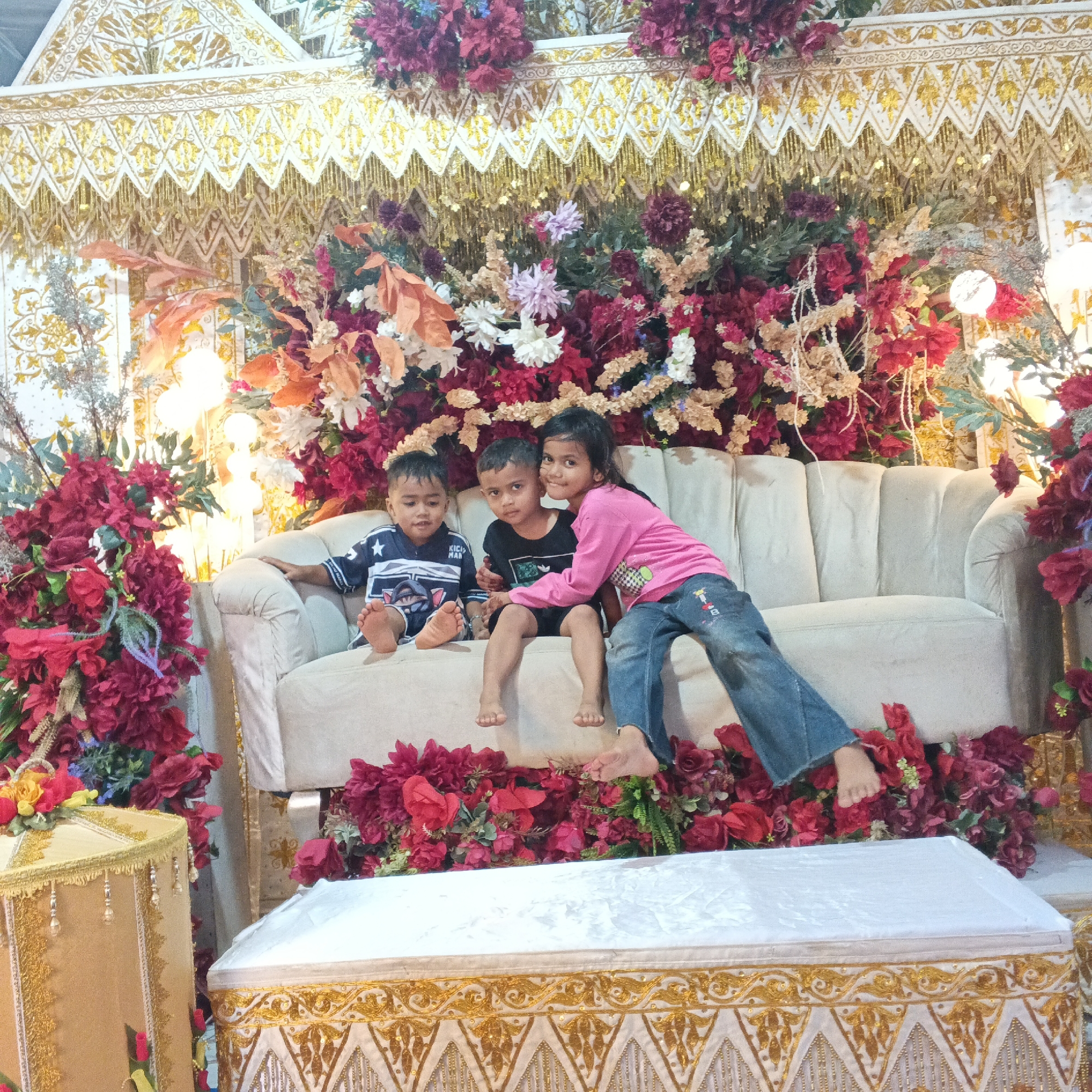 The image depicts three young children posing for a photograph on a plush cream-colored sofa. The children are seated closely together, with a girl on the right embracing the two boys beside her. They are situated in front of an elaborate, ornate backdrop composed of vibrant floral arrangements and decorative golden and white fabric. The backdrop features tiered layers of red and pink flowers, interspersed with lighter-colored foliage and decorative strings. To the left and right of the sofa, decorative boxes or pedestals with gold trim and embellishments are visible, further enhancing the festive and decorated atmosphere of the scene. The lighting appears to be artificial, creating a warm and inviting ambiance. The children are casually dressed, with the boy on the left wearing a navy blue shirt with white stripes and a star graphic, and the boy in the middle in a black t-shirt. The girl on the right is wearing a pink long-sleeved shirt and blue jeans. Their bare feet are visible, resting on the sofa and in front of them amidst the floral decorations. The overall setting suggests a celebratory event, possibly a party or a traditional gathering, given the rich decorations and the posed nature of the photograph.