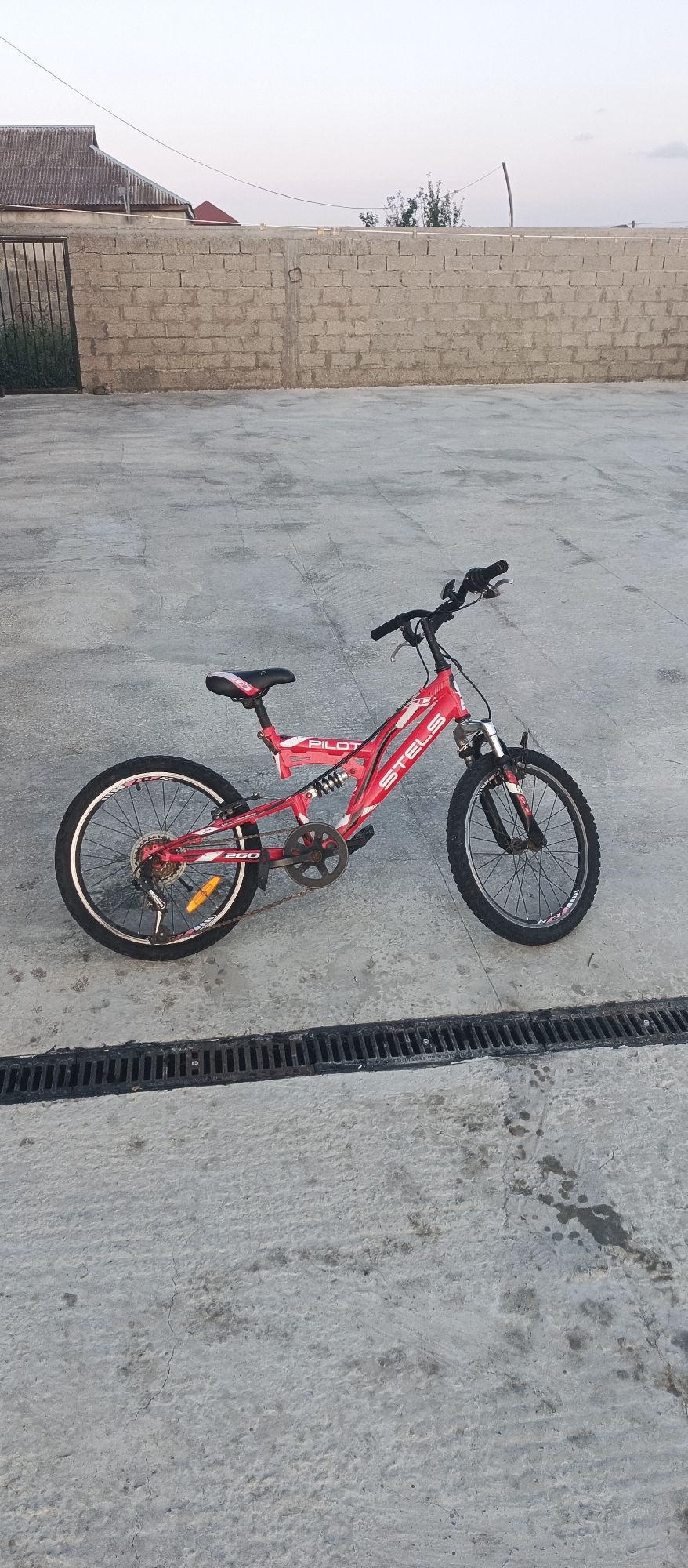 A red and white Stels Pilot bicycle is parked on a concrete surface. The bicycle has a front suspension fork, dual suspension, and 20-inch wheels. The frame is primarily red with white and black accents, including "STELS" and "PILOT" lettering. The bicycle is positioned at an angle, with the rear wheel on the left and the front wheel on the right. In the background, a low, rough brick wall runs horizontally across the frame. Beyond the wall, a residential building with a gray, corrugated roof is visible, along with some trees and a clear sky. The concrete surface in the foreground has a drainage grate. The scene appears to be an outdoor area in a residential or semi-urban setting, possibly a courtyard or driveway.