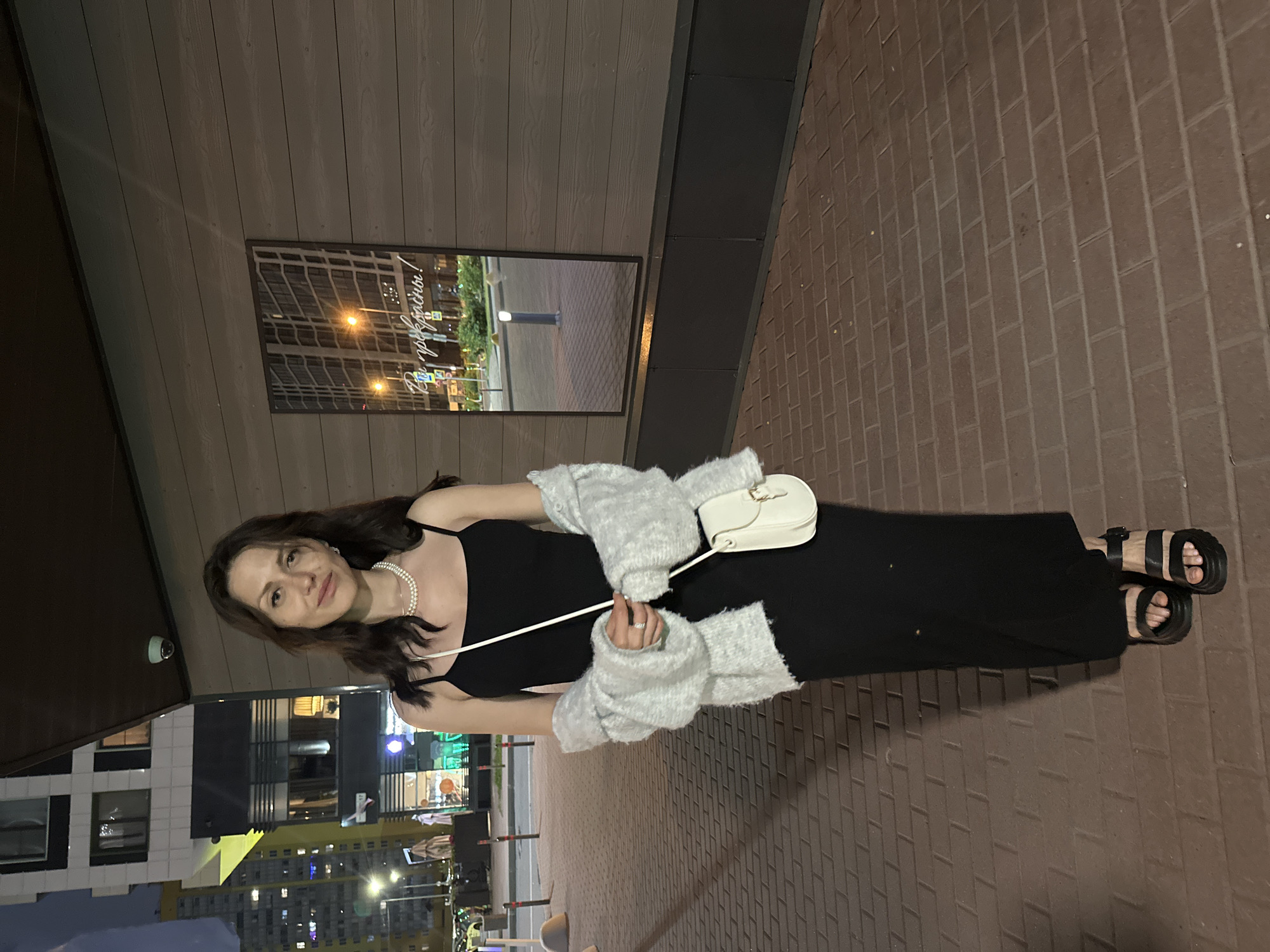 The image depicts a full-body portrait of a young adult female standing outdoors at night in an urban setting in the city of Unknown, Unknown. She stands on a reddish-brown brick-patterned paved surface, slightly angled towards the viewer's right. The subject has long, dark, wavy hair and is looking directly at the camera with a subtle smile. She wears a black, spaghetti-strap midi-length dress, a double-strand white pearl necklace, and a ring on her left ring finger. A light grey, fuzzy cardigan or sweater is draped over her shoulders, held by her arms. She carries a small, off-white crossbody bag with a gold-colored buckle, held in her right hand. Her footwear consists of black platform sandals with two wide straps and an ankle strap.
Behind her, to the left, are multi-story buildings with illuminated windows, suggesting residential or commercial structures. One building is predominantly white with black accents, and another, further back, appears yellow. Streetlights are visible in the background, along with what appear to be street bollards or barriers on the paved area. To her right, a wall composed of horizontal dark wooden or composite panels features a vertically oriented rectangular mirror or reflective panel. The mirror reflects streetlights, buildings, and traffic signs. Cyrillic text, "Вы прекрасны!", meaning "You are beautiful!", is written on the mirror's surface. Below the wooden paneling is a dark grey or black kick panel. A small, light-colored security camera is mounted on the overhang above the wall. The sky is dark, indicating evening or nighttime.