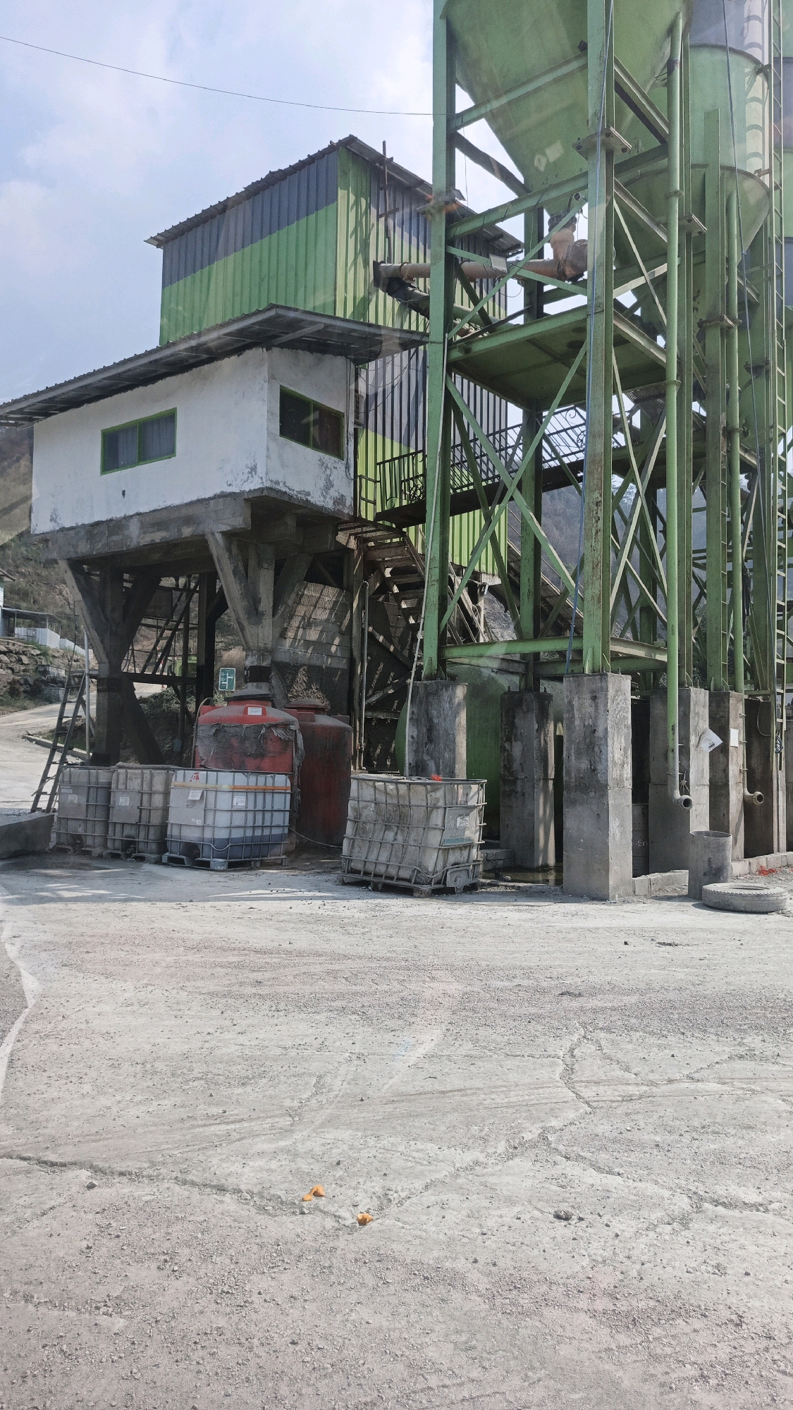 The image depicts an outdoor industrial setting, likely a construction materials processing plant or quarry, in Cijangkar, Indonesia. In the foreground, a rough, gravelly, and paved ground surface dominates, marked with tire tracks and some cracks. Two small orange cones are visible on the ground.
The central focus of the image is a large, multi-story industrial structure. A prominent green metal framework supports large, conical silos. Adjacent to this, a building with a white lower section and a green and black corrugated metal upper section is elevated on concrete stilts. A metal staircase leads up to this elevated building, and a smaller, white structure with green-framed windows is attached to its side.
Beneath the elevated buildings and adjacent to the green structure are several large, rectangular white containers with metal cages, and two large red cylindrical water tanks. A metal ladder leans against the elevated building. The background shows a hillside with some sparse vegetation. The sky is partly cloudy. The overall impression is one of an active industrial site with materials and equipment stored and processed.