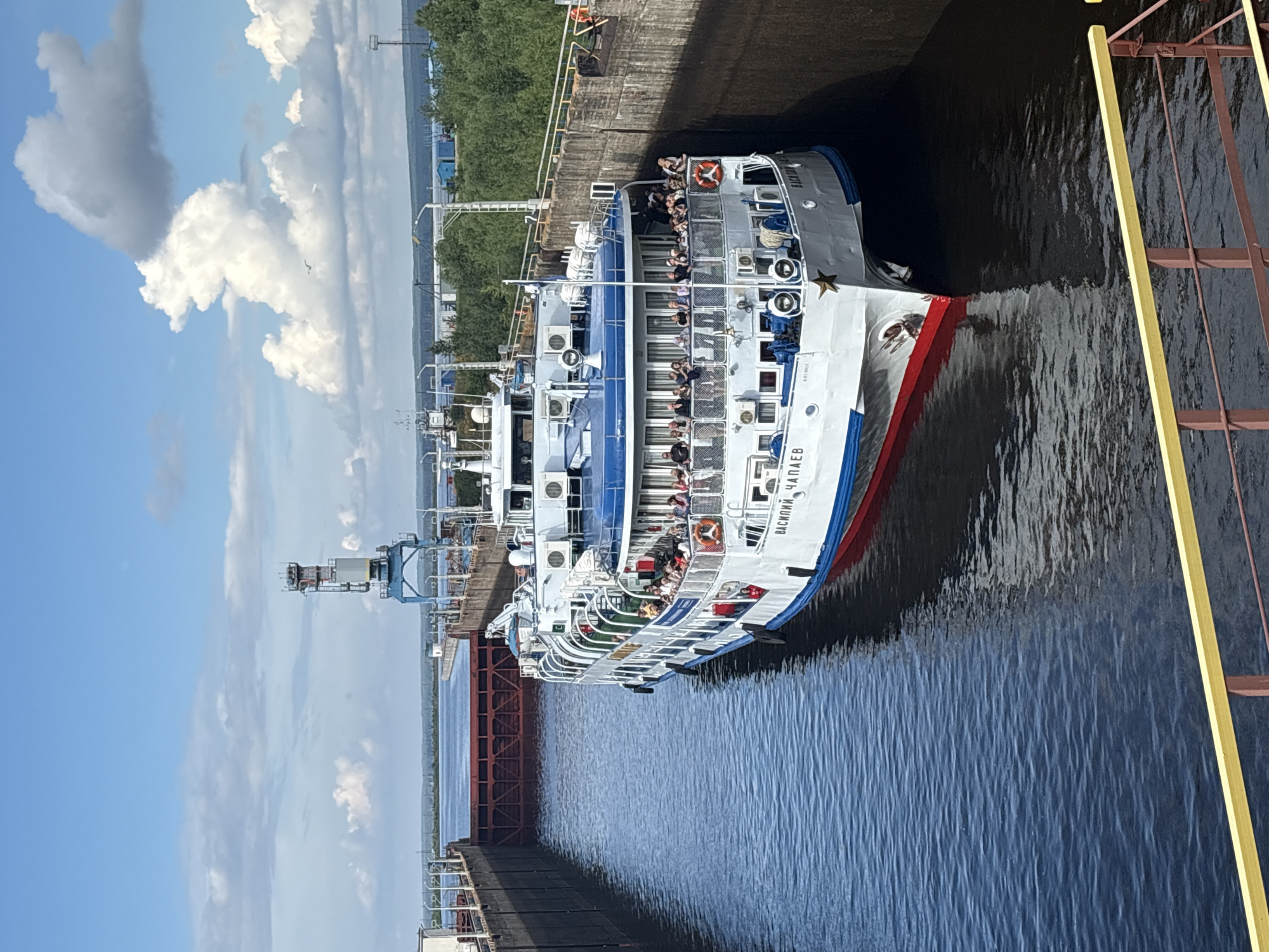 A cruise ship is navigating through a canal lock. The ship is white with blue trim and a red stripe along the waterline. It is filled with passengers enjoying the journey. The canal walls are made of concrete, and a rusty metal structure is visible on the left side of the frame. A yellow railing is on the right side. The sky is blue with fluffy white clouds, suggesting a pleasant day. The water is dark and calm. Text on the ship reads "ВАСИЛИЙ ЧАПАЕВ". The scene likely takes place in Chaikovsky, Russia, given the location context.
