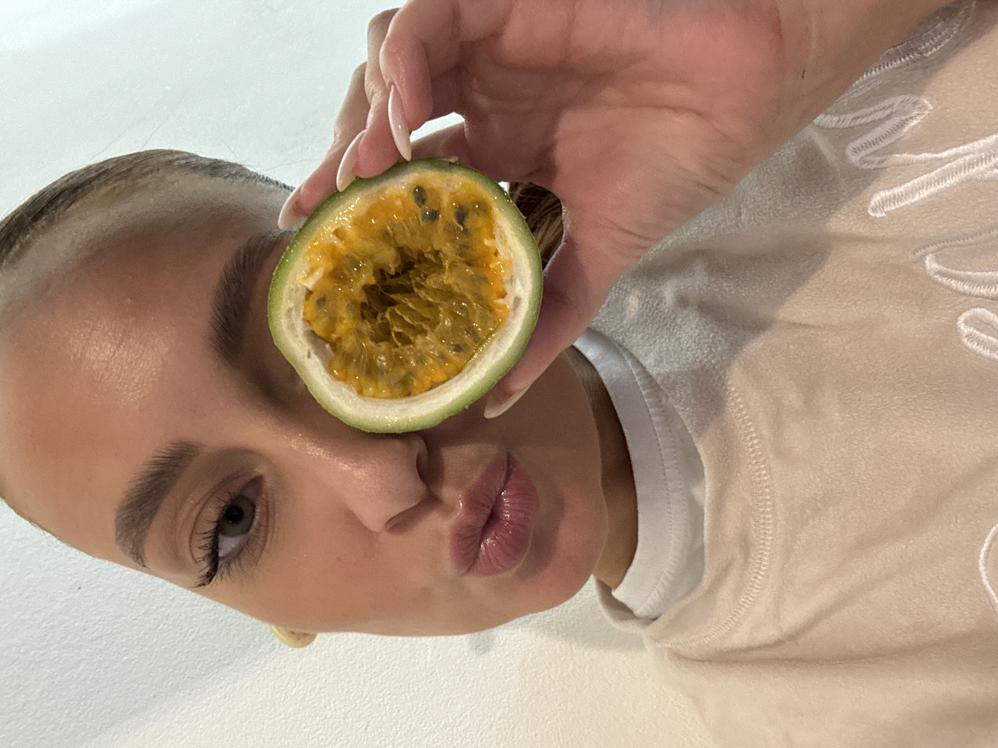 A close-up, selfie-style photograph captures a female individual from the chest up, positioned against a plain, light-colored textured wall. Her hair is pulled back, revealing a clear forehead. The individual's right eye is open, with dark eyelashes and a light-colored iris. Her lips are pursed forward in an expressive pose. She is wearing a light beige or cream-colored fuzzy sweater over a white crew-neck t-shirt. A small, light-colored object is visible near her right ear. Her left hand, featuring long, light-pink manicured nails, holds a freshly halved passion fruit directly in front of her left eye, obscuring that portion of her face. The passion fruit displays its vibrant orange-yellow pulp and black seeds. The scene is evenly and brightly lit, indicative of an indoor setting in the city of Chaikovsky, Russia.