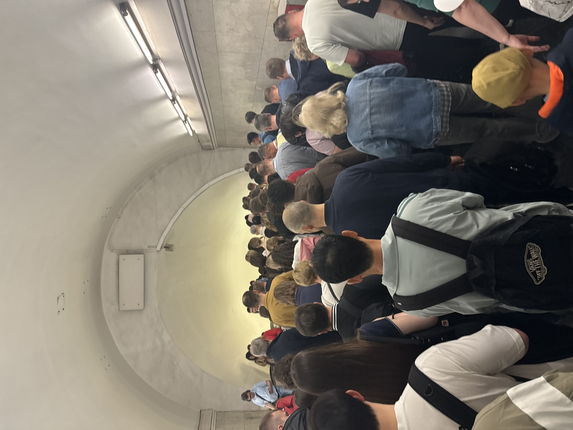 A crowd of people is shown in a subway tunnel, presumably in Moscow, Russia, waiting in a crowded station. The tunnel has a curved ceiling with overhead fluorescent lighting. Most of the people are standing, and some are carrying backpacks or bags. They are dressed in casual clothing.