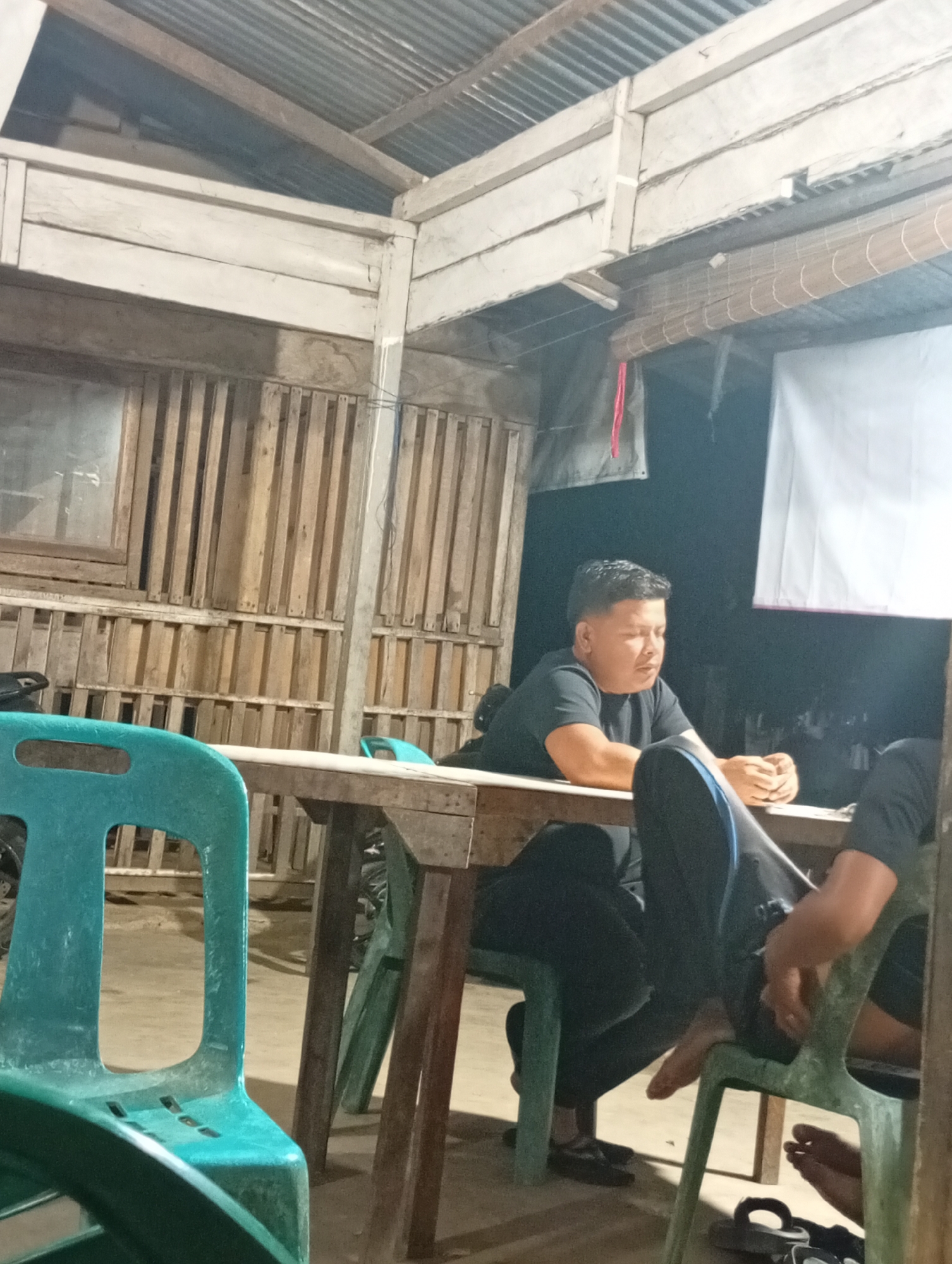 The image shows two men seated at a wooden table, likely in an outdoor, partially covered setting in Lhokseumawe, Indonesia. The man on the left is wearing a black t-shirt and black pants. He is looking down and appears to be resting his chin on his hands, which are clasped on the table. The man on the right is also wearing black clothing, with visible blue stripes on his pants. He is also looking down, with his hands resting on the table. Two turquoise plastic chairs are visible in the foreground and mid-ground, one partially obscuring the man on the left. Behind the men, there is a wall constructed of vertical wooden slats, and above that, a corrugated metal roof is visible. A white banner or cloth is hanging on the dark background to the right of the men. The overall impression is of a casual, informal gathering or meeting.
