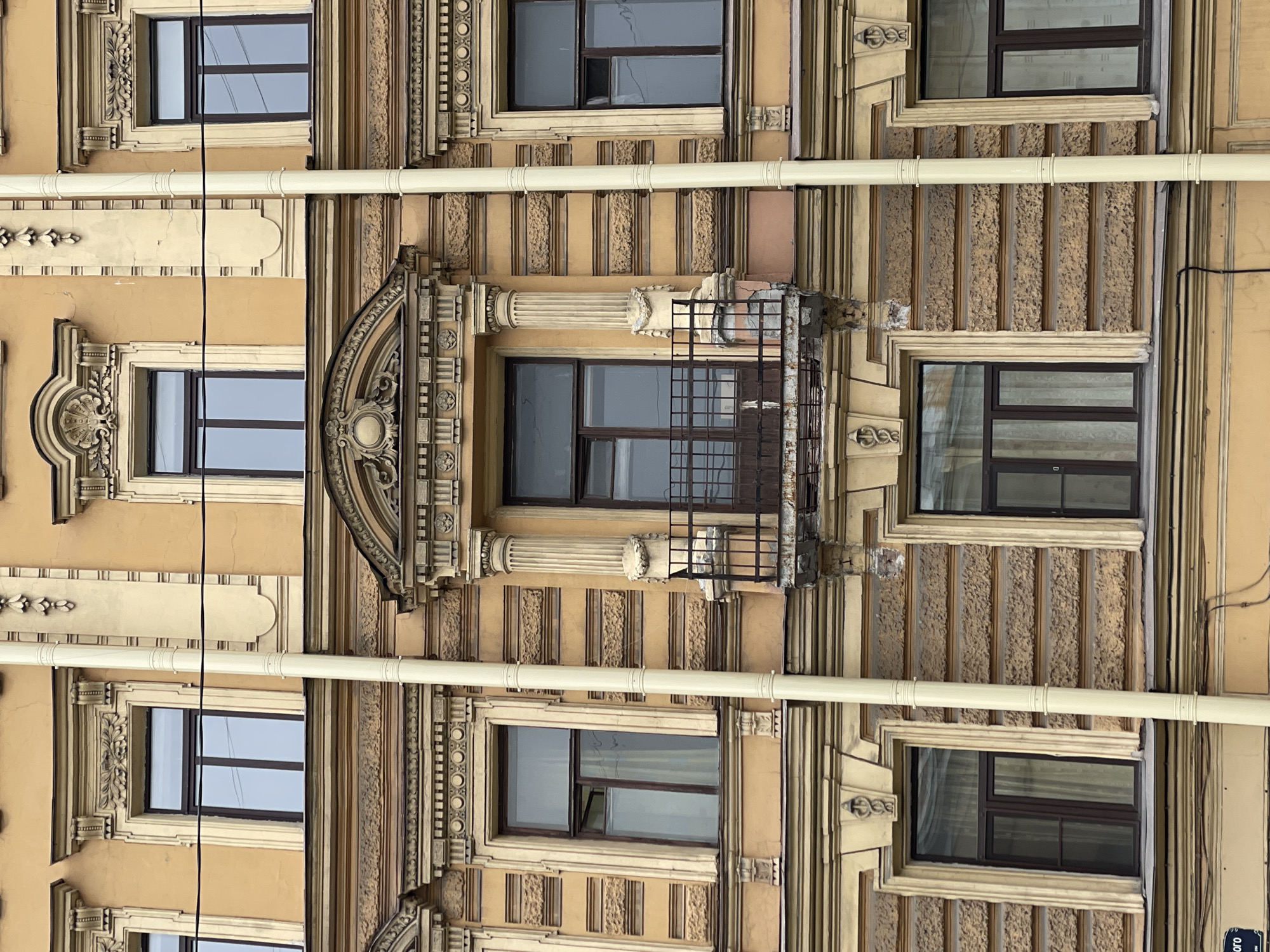 A low-angle, vertical-oriented medium shot features the facade of a building with architectural details. The color palette is dominated by beige, tan, and brown hues. The facade displays multiple windows with dark frames, set within ornately decorated frames featuring raised patterns and intricate carvings.
A distinct architectural element is a decorated balcony, enclosed by a black metal railing. Beige pipes run horizontally across the facade, juxtaposed against the decorative vertical stripes.
The image likely depicts a building in Saint Petersburg, Russia. The architectural style reflects the historical building designs of the city.