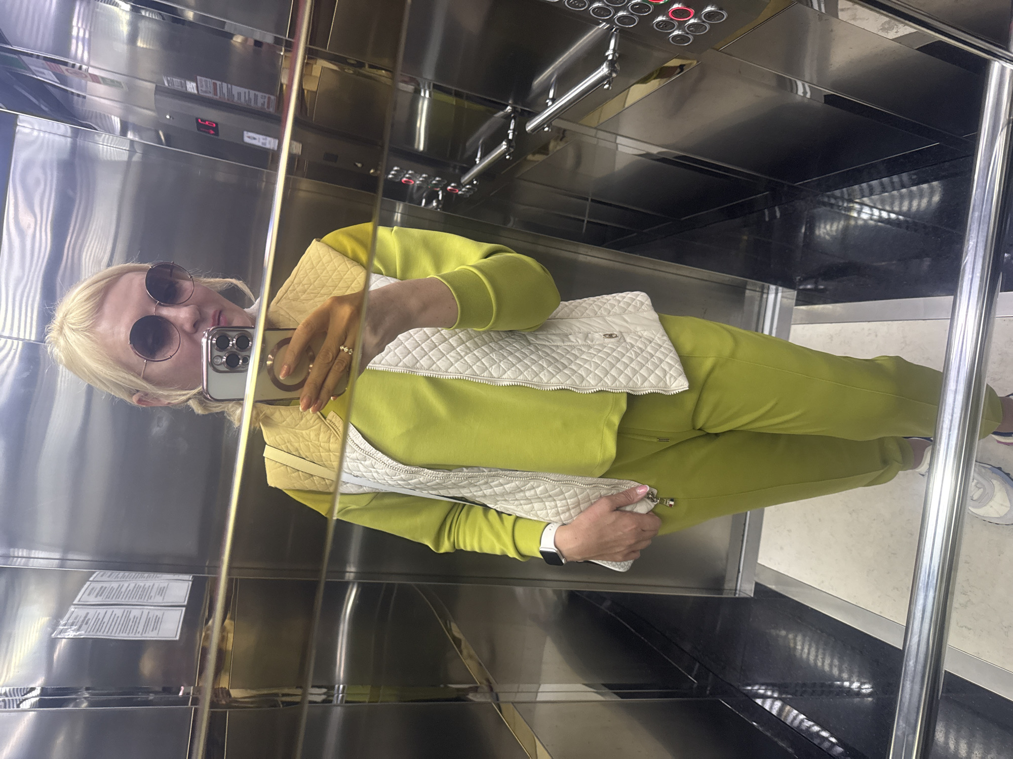 A blonde-haired individual wearing round sunglasses is taking a mirror selfie inside an elevator. The person is dressed in a vibrant lime green tracksuit, layered with a light, possibly off-white, quilted vest. A white smartwatch is visible on their left wrist, and a ring is on their right ring finger. They are holding a silver smartphone, likely an iPhone, in their right hand, angled towards the mirror to capture the reflection. A white quilted bag or clutch is also visible, held in their left hand's reflection.
The elevator interior features highly reflective, polished metal walls, some appearing silver and others with a golden or brass-like tint due to reflections. A control panel on the right wall contains multiple round buttons, several of which are illuminated in red. A small digital display on the panel shows red characters, possibly "LO" or "40," next to an arrow, indicating the current floor or direction. A metal handrail runs horizontally along the wall. The floor of the elevator is a light beige or cream color. On the left wall, two rectangular white signs with dense, small black text are affixed, though the text is illegible due to resolution and reflections. The overall scene depicts a casual moment, possibly a quick outfit check or social media post.