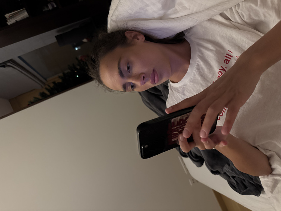 A person is lying in bed, engrossed in their phone. They're wearing a white t-shirt, their hair pulled back. The phone is held in both hands, fingers poised to tap the screen. The room is dimly lit, with a glimpse of a shelf and greenery visible in the background. Bedclothes are rumpled, suggesting comfort and relaxation. The focus is on the person's concentration as they interact with their device.