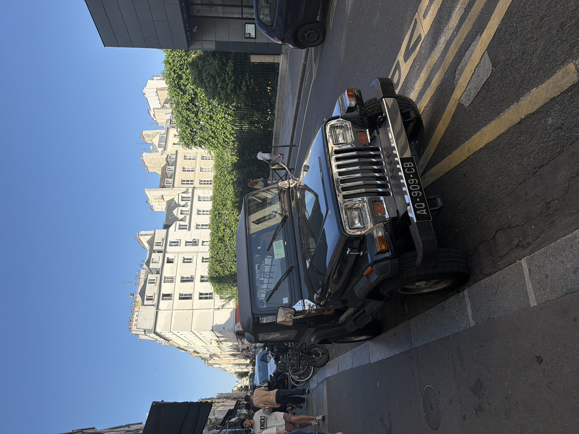 The image shows a black Jeep Wrangler parked on a street in Paris, France. The car is oriented facing the upper right of the frame, with its front license plate visible, reading "AQ-909-CB." The surrounding environment consists of buildings with Parisian architecture, trees, parked bicycles, and other parked vehicles. People can be seen walking on the sidewalk. The image appears to have been taken during the daytime, with a clear, blue sky visible. Yellow painted stripes can be seen on the asphalt.