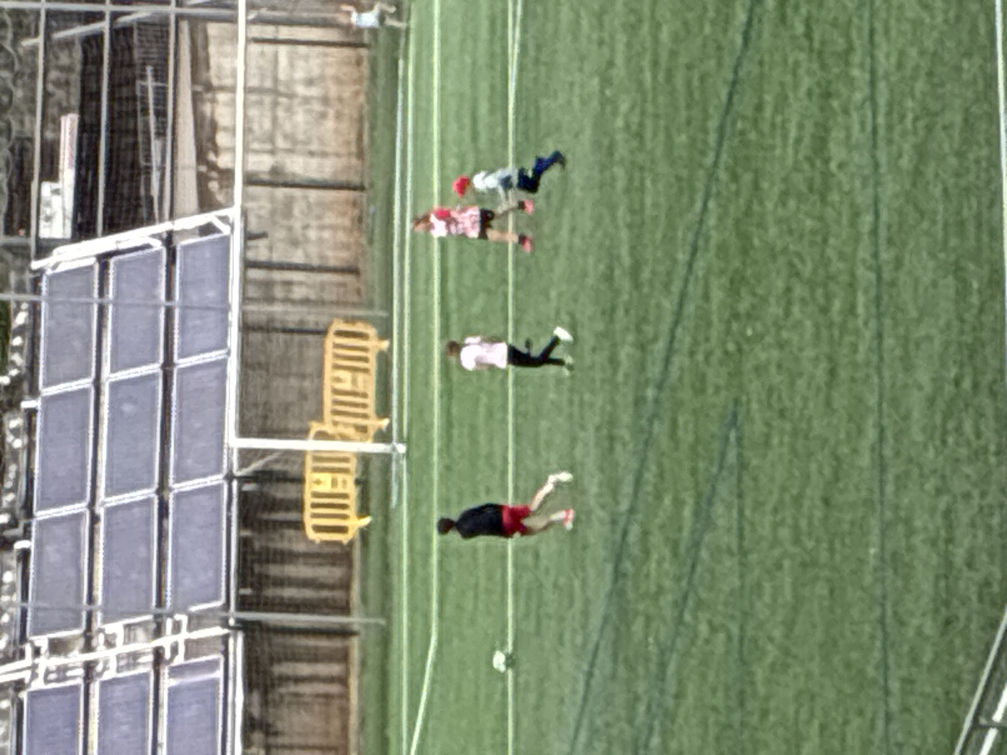 The image shows a soccer field with four people playing soccer. The field is green with white lines. A soccer ball is on the ground near one of the players. To the left of the field is a building with barred windows. The photo was taken in Esplugues de Llobregat, Spain.