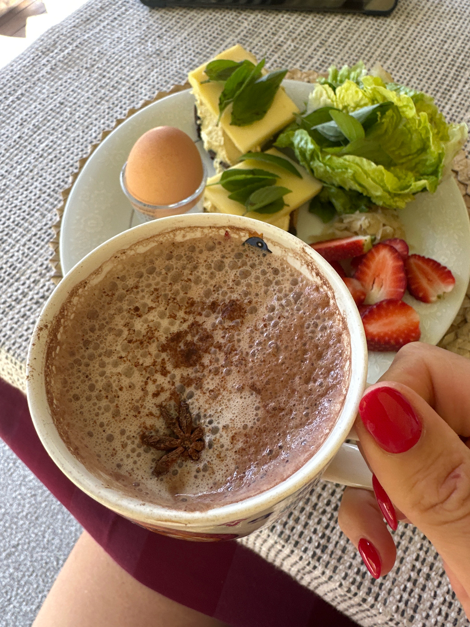 A close-up overhead shot shows a cup of hot chocolate with star anise and cinnamon on top, placed on a table with a textured tablecloth. Next to it, a white plate holds a boiled egg in a cup, slices of cheese with mint leaves, lettuce, and sliced strawberries. A hand with bright red nail polish is holding the cup. The setting appears to be a casual meal, possibly breakfast, with a focus on food and drink. The image does not provide any information about the city of Thora, Australia or any specific location within it.