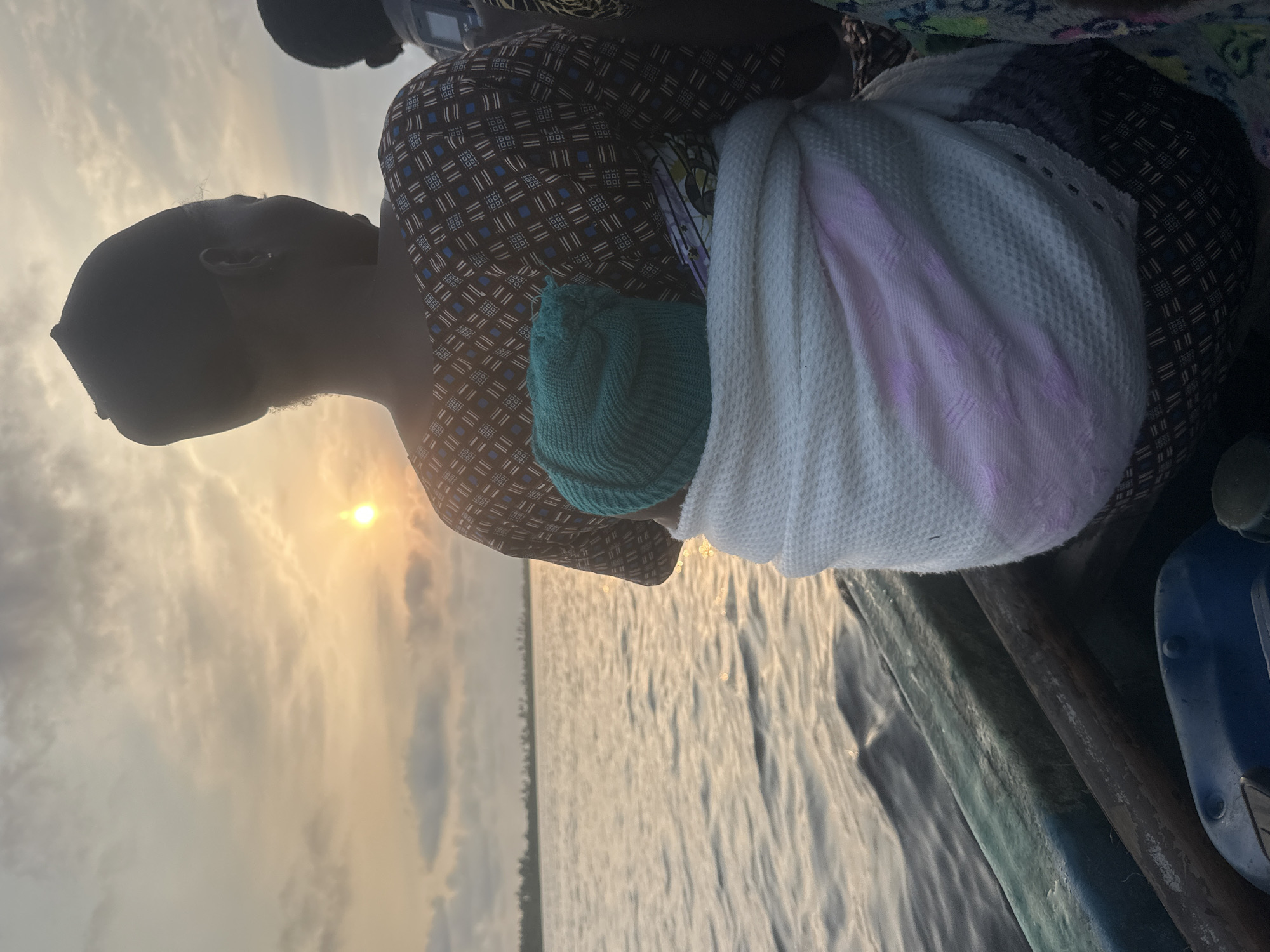The image depicts a rear view of a woman and a baby in a boat on a body of water, likely during sunset. The woman, wearing a patterned top and a dark head covering, is carrying a baby wrapped in a white and pink blanket, with the baby wearing a teal knitted hat. The boat is wooden, with visible oar handles and a blue container in the foreground. In the background, a tree line is visible along the horizon, and the sun is setting, casting a warm glow on the water and sky, suggesting the location is coastal. The overall atmosphere is serene, captured from a low angle as the boat moves through the water. This scene is consistent with locations around Badagry, Nigeria, known for its waterways and coastal scenery, especially during twilight hours.