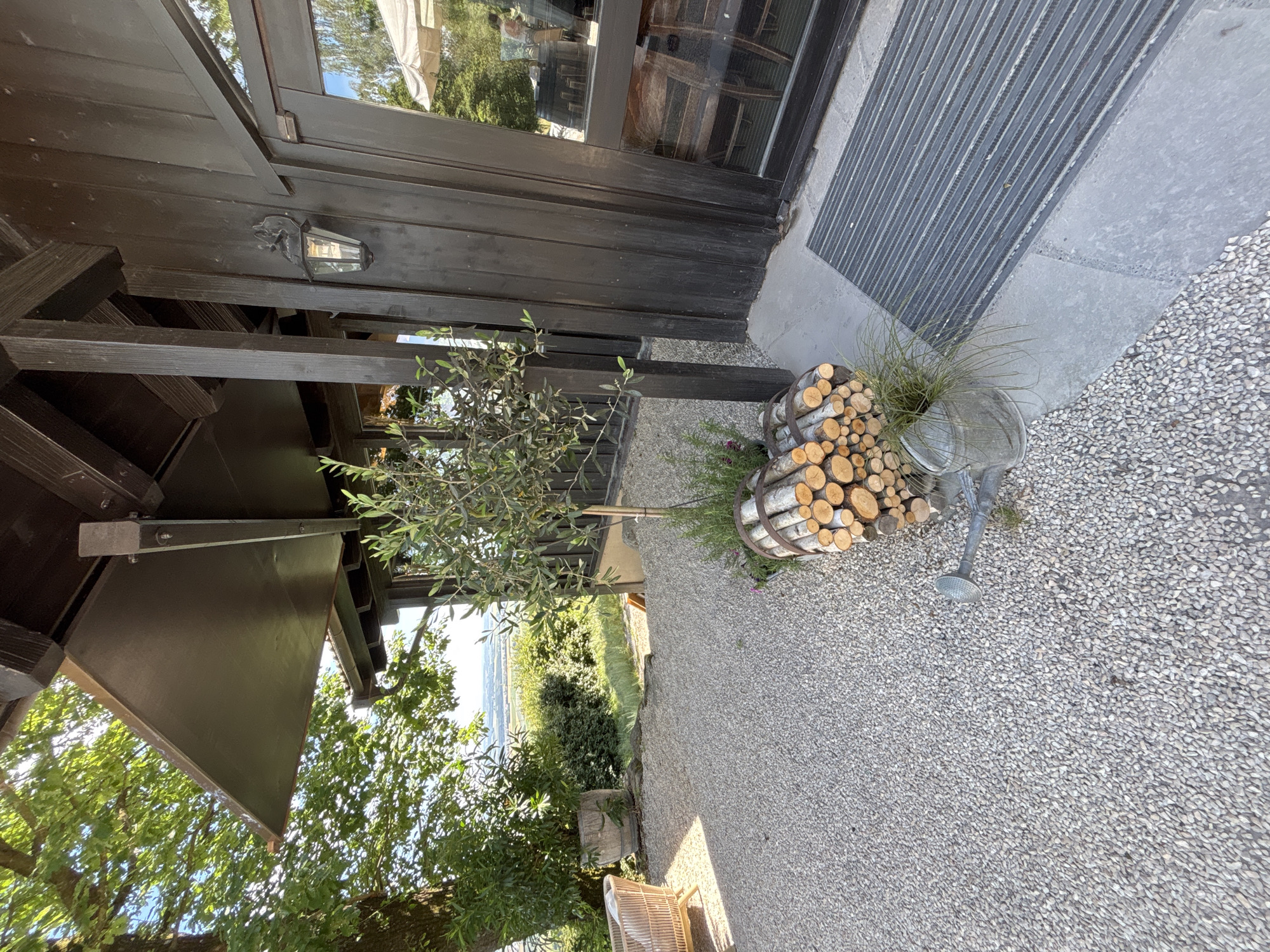 A low-angle, eye-level outdoor shot captures the exterior of a dark wood building with a covered patio area. The building has a pitched roof supported by dark wooden beams and a large window with reflections of trees and an outdoor seating area. To the left, a large tree shades a portion of a light-colored gravel patio, where a woven chair is visible. A large wooden barrel plant pot sits near the tree. In front of the building, near a stack of firewood and a watering can, a small olive tree is planted. A grey doormat is positioned in front of the entrance. The background reveals a scenic view of rolling hills and vegetation under a bright, sunny sky.