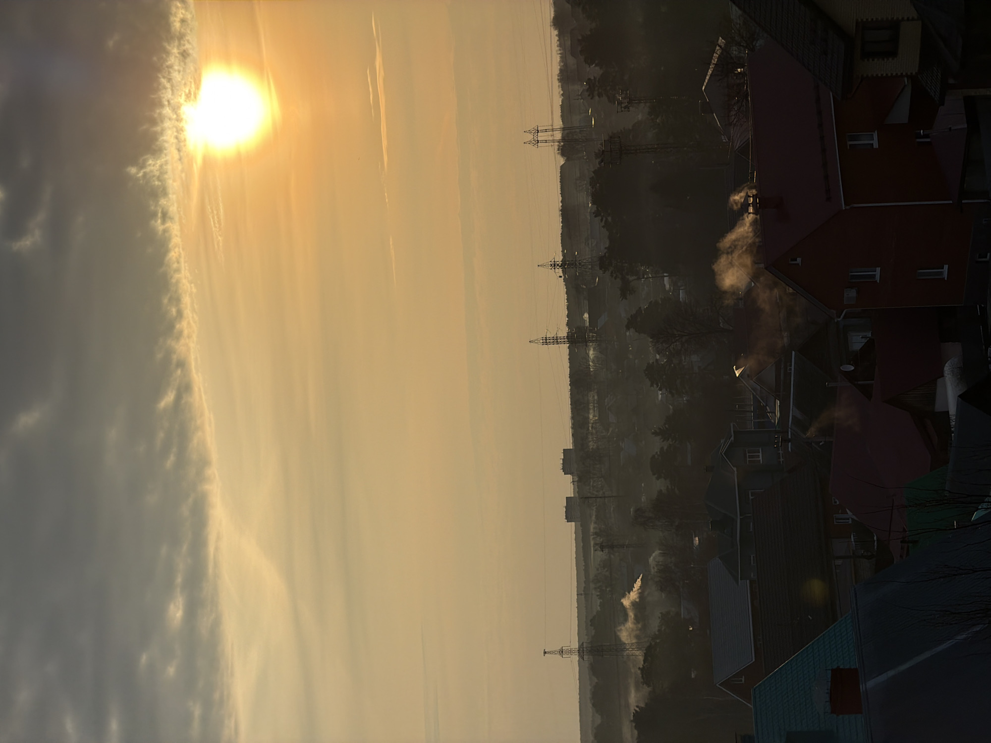 The image depicts a cityscape at sunrise or sunset, likely in the city of Chaikovsky, Russia. The sun is prominent on the left, partially obscured by clouds, casting an orange-yellow hue across the sky. Power lines stretch across the central part of the scene, leading into a slightly hazy and wooded urban area. Smoke rises from chimney stacks of the houses. The houses are mostly built with brick, some houses have red roofs. No people are visible in the image.