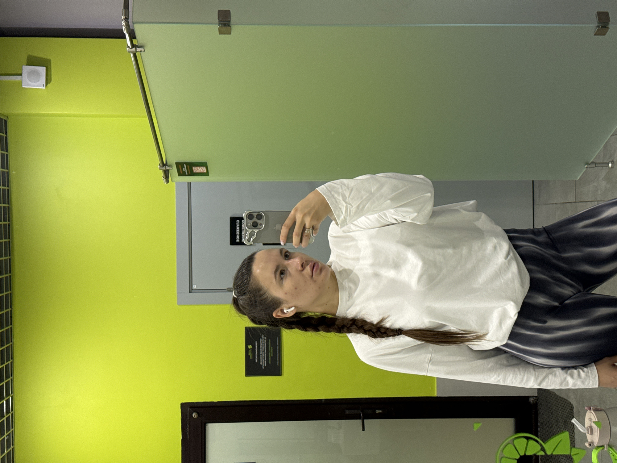 A woman is captured in a selfie reflection, holding a smartphone, likely an iPhone, in front of a bright lime green wall and a translucent light green partition. She has light brown hair tied back with a single braid extending over her shoulder, and is wearing white wireless earbuds. Her expression is a slight pout. She is dressed in a loose-fitting white long-sleeved top and dark blue or black pants with lighter horizontal stripes. A ring is visible on a finger of the hand holding the phone.
The background features a vibrant lime green wall on the left, with a white rectangular speaker or sensor mounted on it. A grey utility door or cabinet is visible behind the woman, partially obscured by her body and the phone. Above and to the right, a frosted or translucent light green partition extends across the frame, held by metallic fixtures. On this partition, a small green plaque with reversed, unreadable text is visible. Below the partition, on the lime green wall, there is a dark-framed sign with white text that appears to be in a non-English language (possibly Russian, given the location context), which is also mostly unreadable due to distance and angle. Part of a dark door frame is visible at the bottom left of the image. The setting appears to be an indoor public space, possibly a gym or changing room, characterized by the bright walls and functional fixtures.
