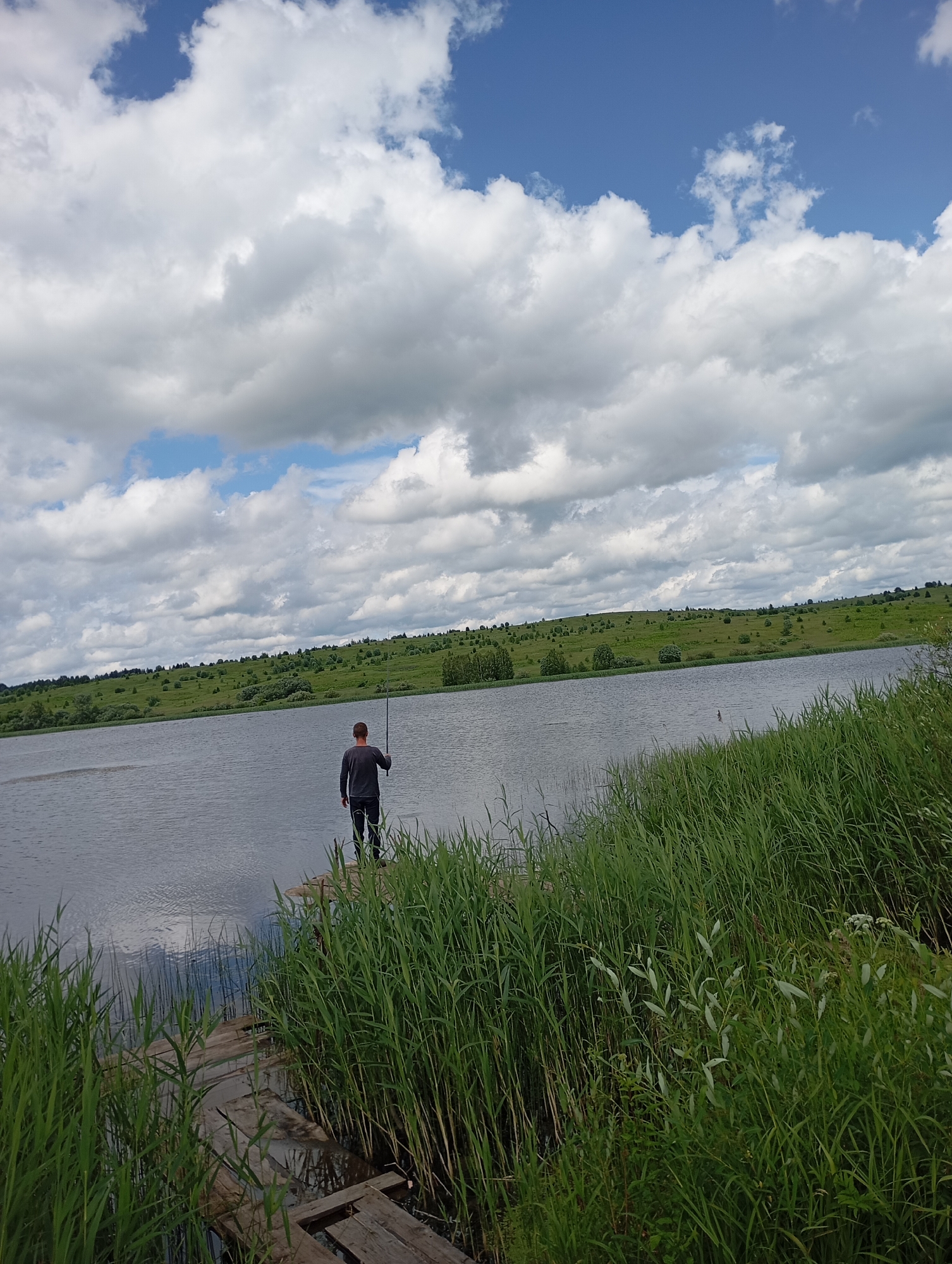 The image depicts a person fishing by a lake in the village of Ryabki, Russia. The individual stands on a wooden pier, facing the water, with a fishing rod in hand. The lake is calm, reflecting the cloudy sky. Lush green reeds and grasses line the shore in the foreground. In the background, rolling hills covered in green vegetation and scattered trees stretch towards the horizon. The sky is partially covered with large, white, cumulus clouds against a backdrop of blue sky.