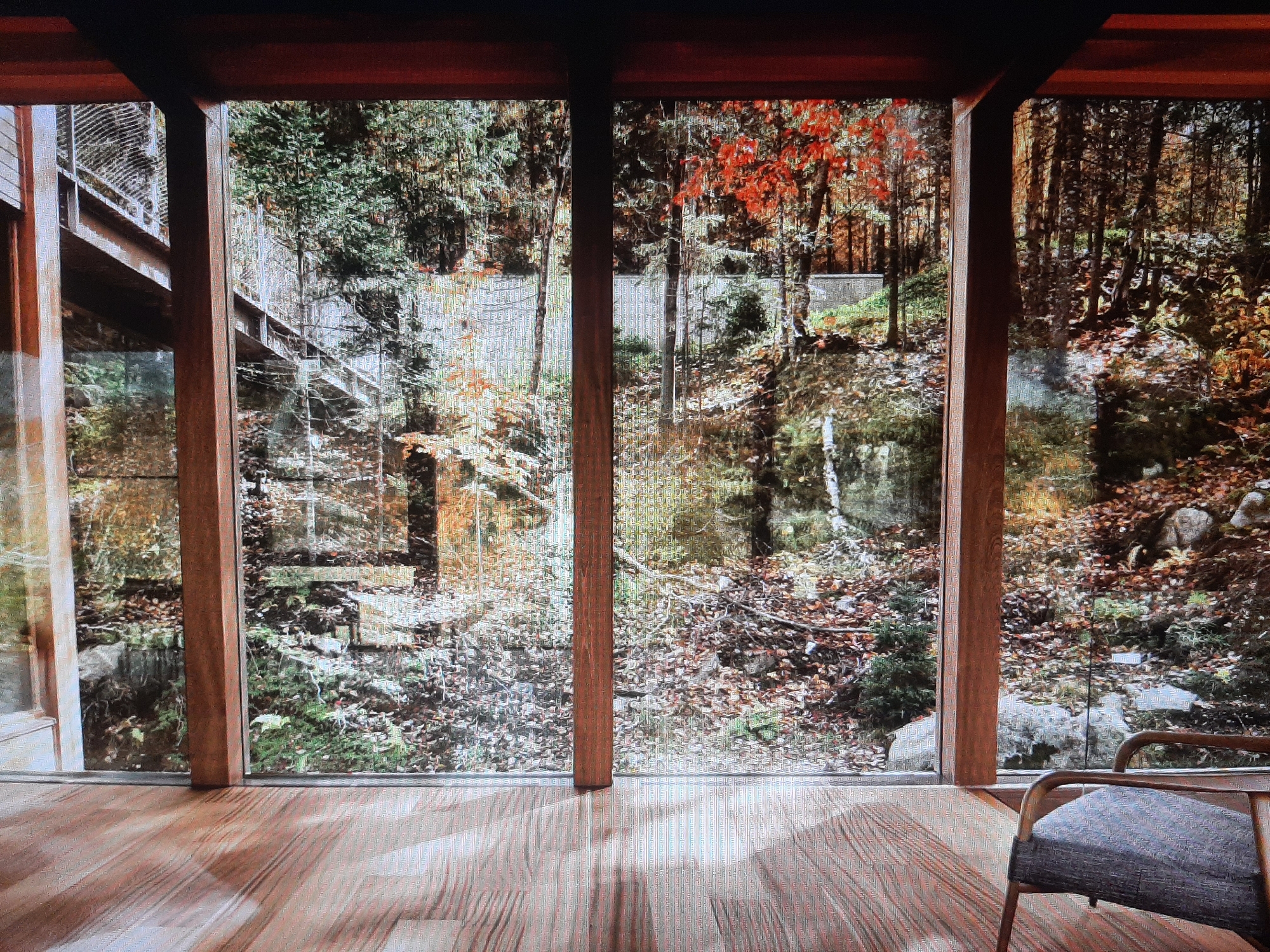 A wide shot captures the interior of a room with a floor made of light-colored wooden planks. The room faces a large window that looks out onto a wooded area with a bridge. The window is divided by three vertical wooden beams. The left side of the window shows a section of the wooden bridge with a metal railing, and the reflection of trees on the glass. The middle and right sections of the window display a dense forest with trees in various stages of autumn foliage, with leaves scattered on the ground. The floorboards show a wood grain pattern and are illuminated by natural light. In the lower right corner of the image, a portion of a modern chair with a dark patterned cushion and metal frame is visible. The overall scene is one of a modern interior with a view of nature.