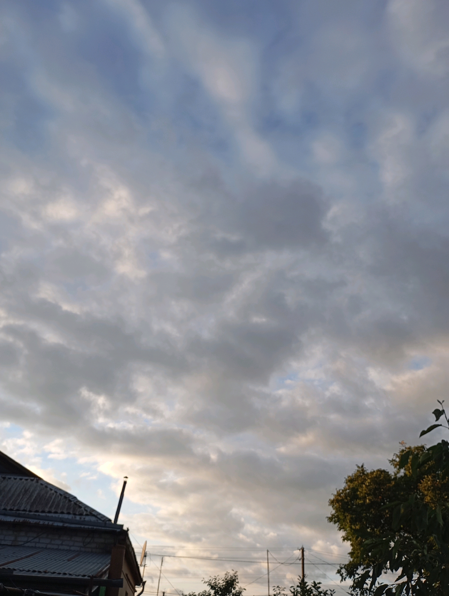 The image depicts a cloudy sky over a residential area in Horishni Plavni, Ukraine. The lower left corner shows a portion of a building with a corrugated metal roof and brick walls. A tall, thin metal pole extends upwards from the roofline. To the right, a large tree with dark green foliage is visible against the sky. Electrical wires and utility poles are also present in the background, suggesting an urban or suburban setting. The clouds are varied in density, with patches of lighter sky visible through them, indicating a partly cloudy or overcast day. No people are visible in the frame.