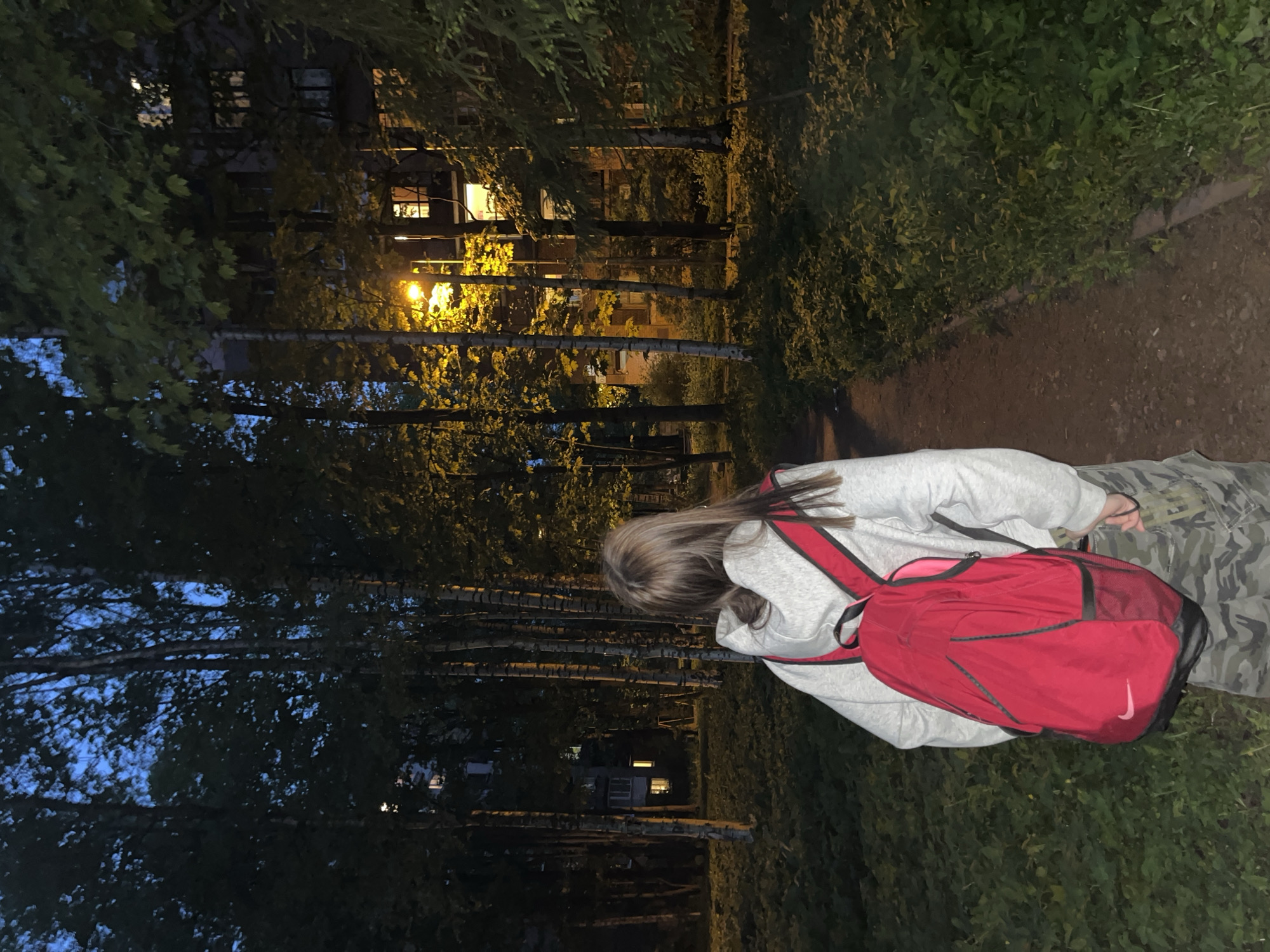 A woman wearing a gray hooded sweatshirt and camouflage pants stands on a dirt path with green foliage to the right. She has a red Nike backpack on her back. To the left, there are tall trees, some with white trunks. Buildings with lit windows are visible through the trees. The scene appears to be in Saint Petersburg, Russia, given the context. It is dusk or night. There is a streetlight visible amongst the trees. The woman is facing away from the camera.