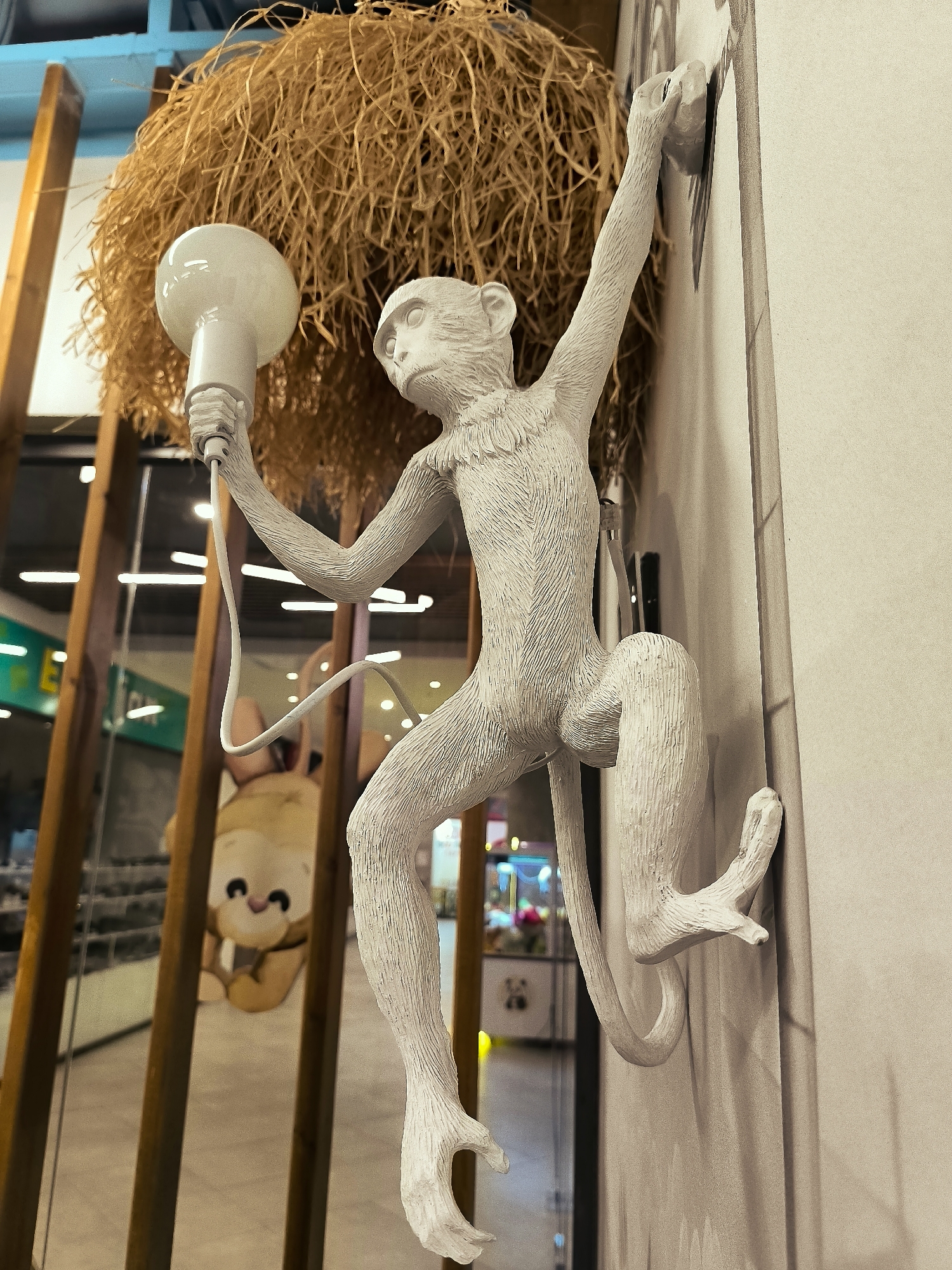 A white, stylized monkey sculpture is mounted on a light-colored wall. The monkey is depicted in a dynamic pose, with its right hand reaching up and its left hand holding a white lightbulb attached to a wire. Its tail curls around its body. Behind the monkey, a large, spherical lampshade made of straw-like material hangs. To the left of the monkey sculpture, a blurry image of a stuffed toy, possibly a bear, is visible through a wooden structure. The background appears to be an indoor setting, with reflections of lights and store displays visible. The scene is indoors and does not depict any specific location within the Qarağandı city administration.