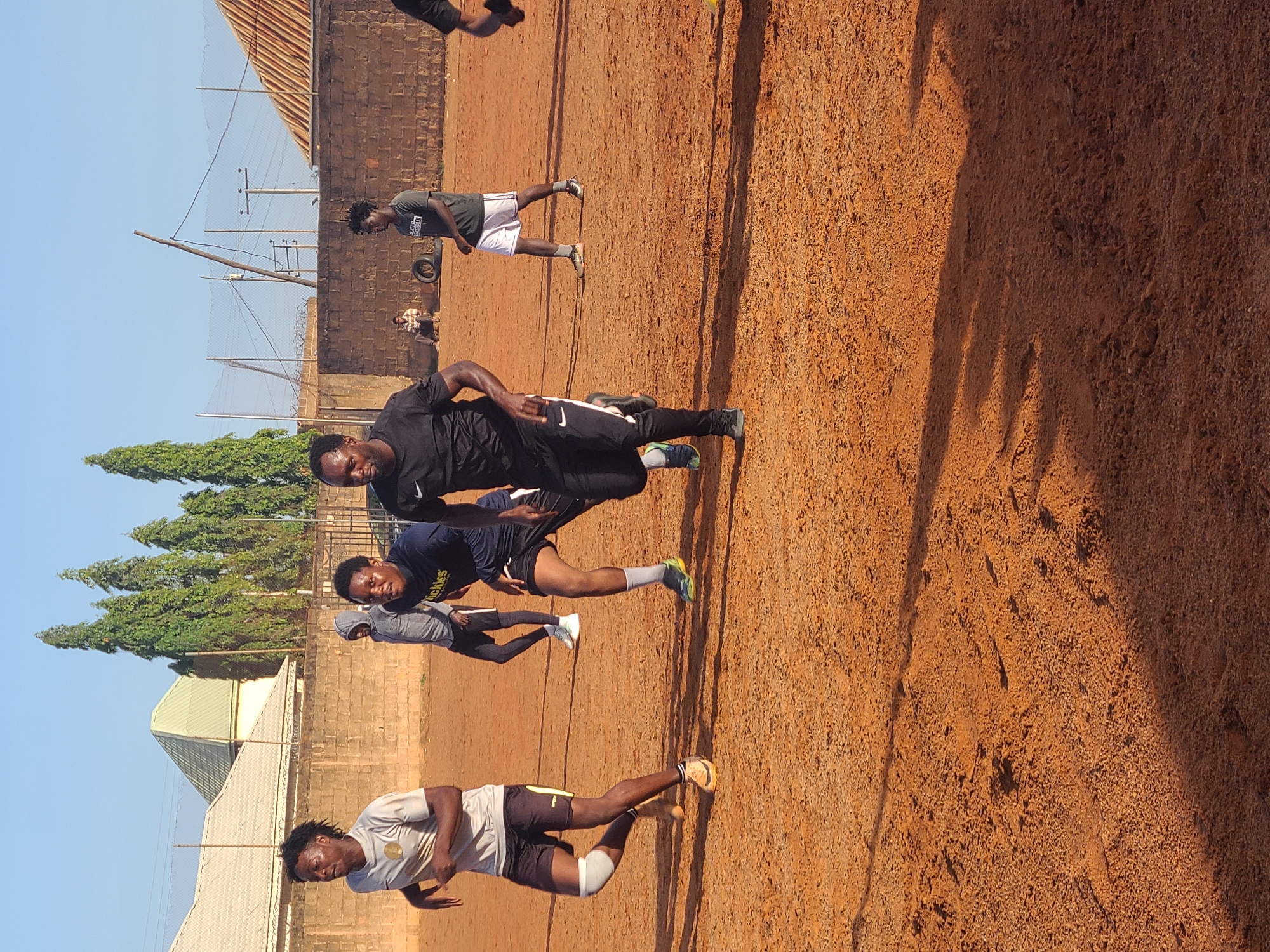 The image displays an outdoor scene, likely a sports or training ground, under a clear blue sky. Several young men are present, engaged in what appears to be physical activity or a break from training. The ground is a reddish-brown dirt or clay, showing variations in texture and footprints. Long, distinct shadows are cast across the ground, indicating a low sun angle, suggesting either morning or late afternoon.
In the foreground, a man wearing a black athletic top and black pants with white stripes stands with his left hand on his hip, looking towards the camera with a slight smile. To his right, another man in a grey athletic shirt with "THE REAL BASEBALL" printed in white, and white shorts, is captured in a dynamic stance. Behind him, a man in a blue athletic shirt, which has yellow text reading "Riches" or "Ritchies," is bending forward. Further in the background, other individuals in athletic wear are visible, including one in a grey hooded top.
The background features a line of tall, green, conical trees on the left side, separating the field from distant buildings, one with a light-colored roof. On the right, a low brick wall topped with a chain-link fence, possibly with barbed wire, borders the field. Beyond this wall, utility poles with wires rise against the sky, and a building with a rusty, corrugated metal roof can be seen. A discarded tire rests on the ground near the wall. The overall atmosphere suggests an informal yet focused outdoor athletic gathering.