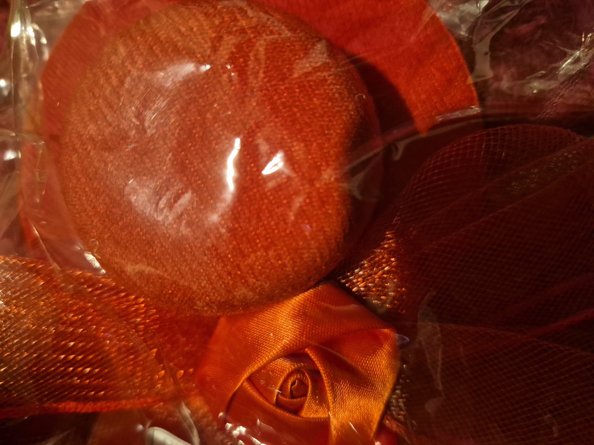 The image shows a close-up view of red decorative items, possibly part of a gift wrapping or craft project, enclosed in clear plastic. In the foreground, a ribbon has been fashioned into a rose shape, its satin petals tightly coiled. To the right of the rose, a round, textured object, also in a vibrant red hue, is visible, its surface appearing slightly granular. Another textured red material, possibly netting or mesh, is layered behind the rose and the round object, creating a visual depth with its criss-cross pattern. The overall impression is one of festive or celebratory embellishments, characterized by rich red tones and varied textures, all contained within a transparent wrapping that diffuses the light and creates a somewhat glossy sheen. There is no indication of any specific location within the city of Sandwell, United Kingdom.