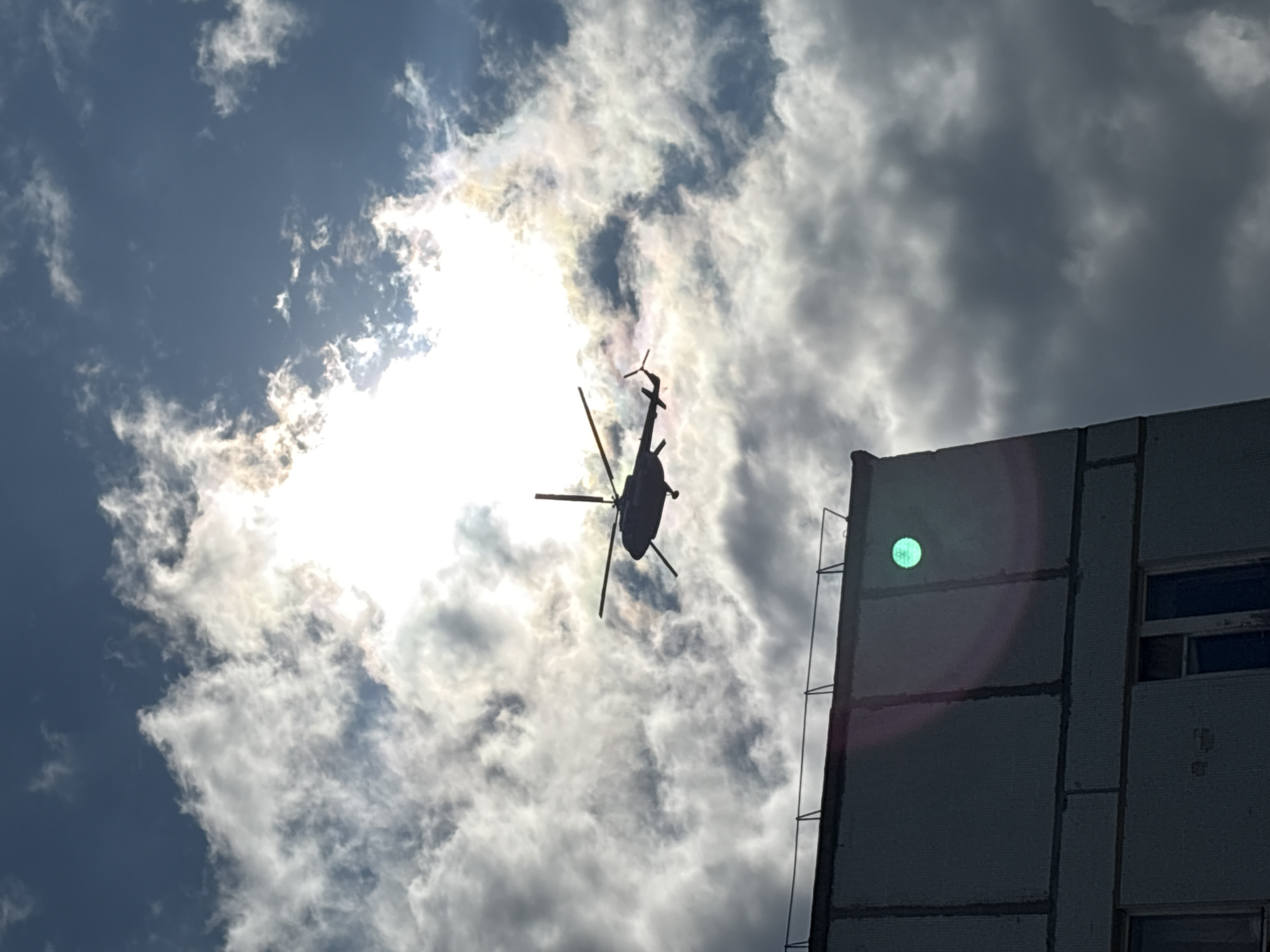 A silhouette of a helicopter is visible in the sky above a building in Chaikovsky, Russia. The helicopter is in mid-flight, with its rotors spinning. The sky is partly cloudy, with the sun shining through the clouds, creating a bright halo effect. The building is a multi-story structure with a concrete facade. A bright green light reflection is visible on the building's wall.