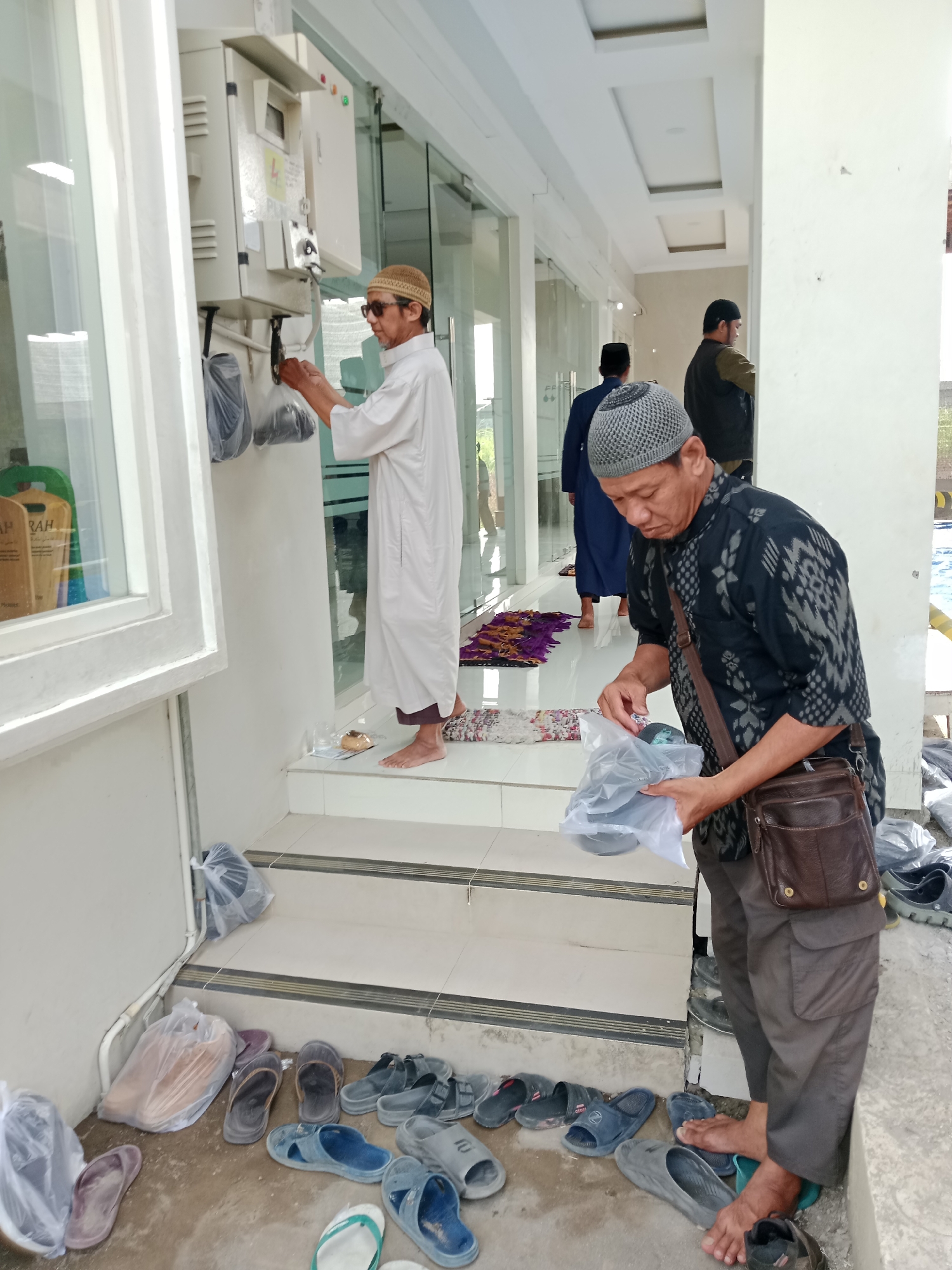 In Kediri City, Indonesia, two men are seen standing outside what appears to be a building with large glass doors and a white facade. The man in the foreground, wearing a grey patterned skullcap and a dark patterned shirt, is holding a plastic bag filled with dark-colored footwear. He has a brown leather satchel slung across his chest. To his left, and further back, a second man in a white robe and a beige skullcap is reaching for a black bag hanging from a metal electrical box mounted on the wall. Behind these two, another man in a dark vest and a black skullcap is visible, facing away from the camera and appearing to be in the process of entering the building. The ground is covered with a variety of sandals and shoes, some in plastic bags, suggesting a place where footwear is removed before entry. Prayer mats, one purple and one multi-colored, are laid out on the tiled steps leading up to the building's entrance. The overall environment suggests a religious or communal gathering space.